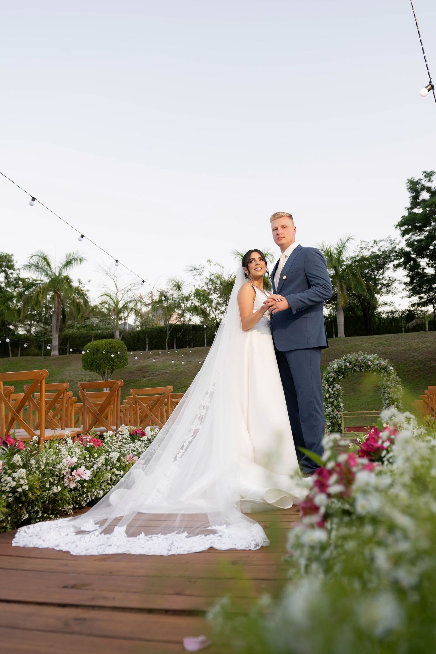 Bride and groom posing on a wooden deck surrounded by flowers at an outdoor wedding venue.