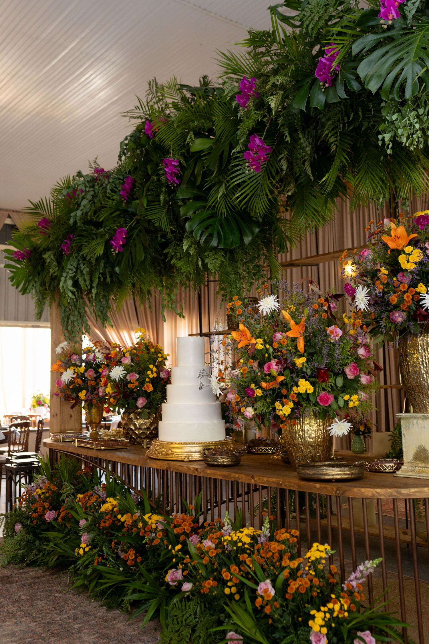 Lush tropical wedding dessert table with white tiered cake, colorful flowers, and vibrant green foliage canopy.