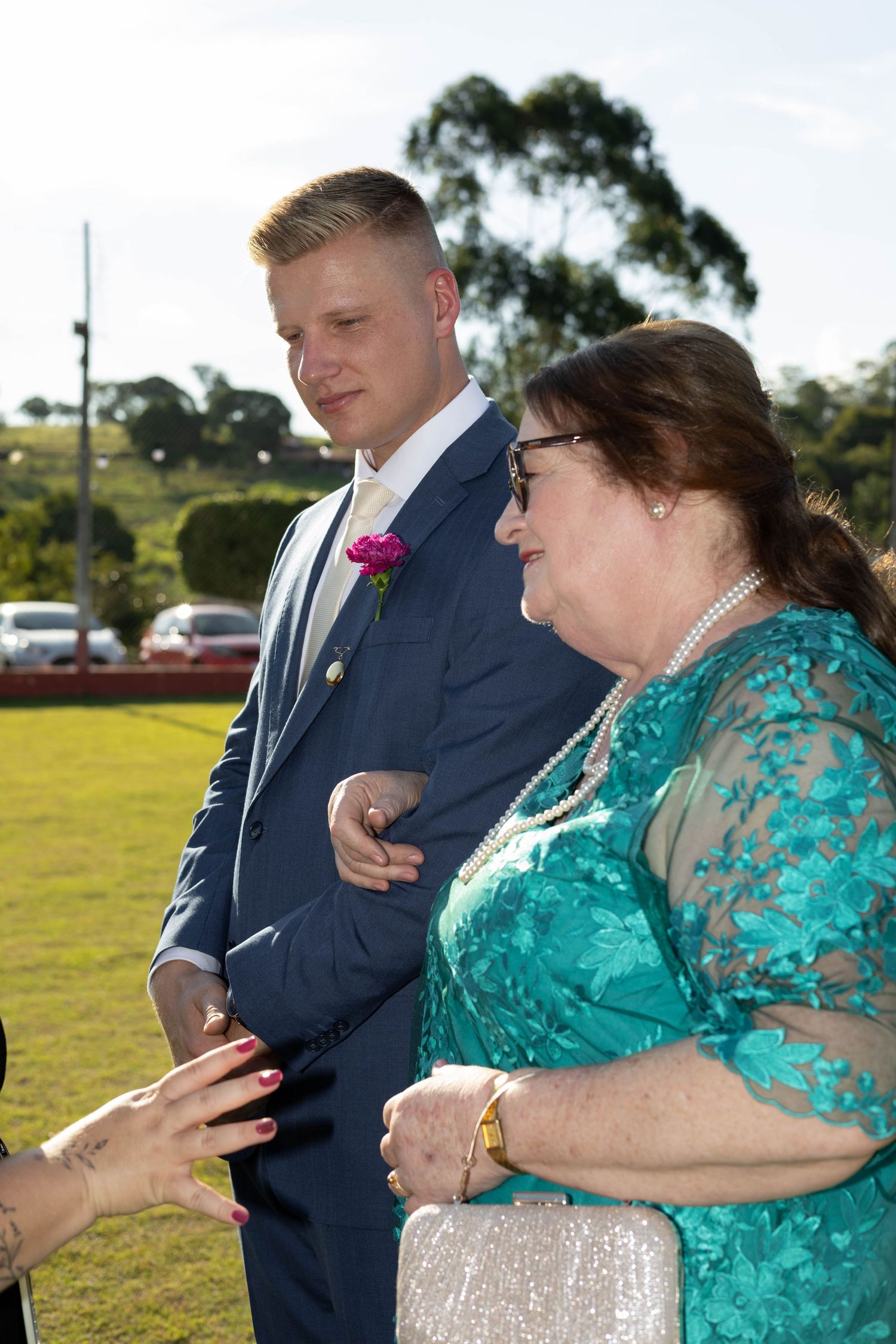 Groom in navy suit with pink boutonniere stands beside woman in teal lace dress at outdoor wedding ceremony.