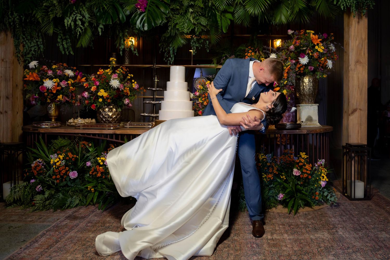 Groom dips bride in romantic kiss at wedding reception with floral backdrop and white tiered cake.