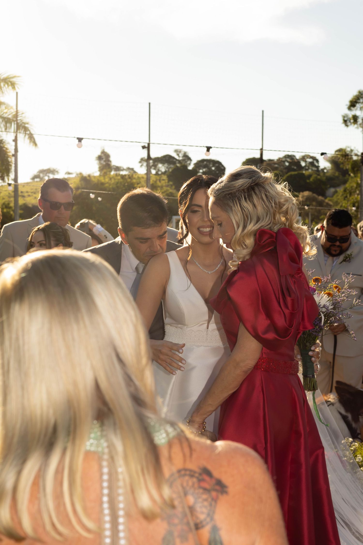 Bride in white dress laughs joyfully with guests in red and gray attire at sunny outdoor wedding.