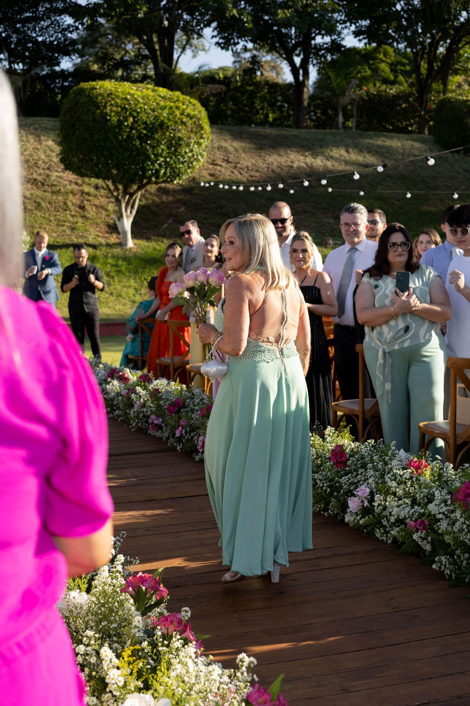 Bridesmaid in sage green gown walks down floral-lined aisle at outdoor wedding ceremony with guests standing.