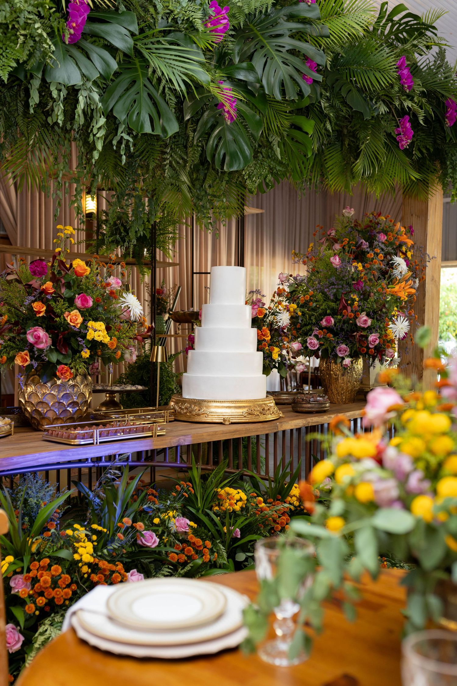 Elegant white tiered wedding cake on gold stand surrounded by colorful tropical floral arrangements and lush greenery.