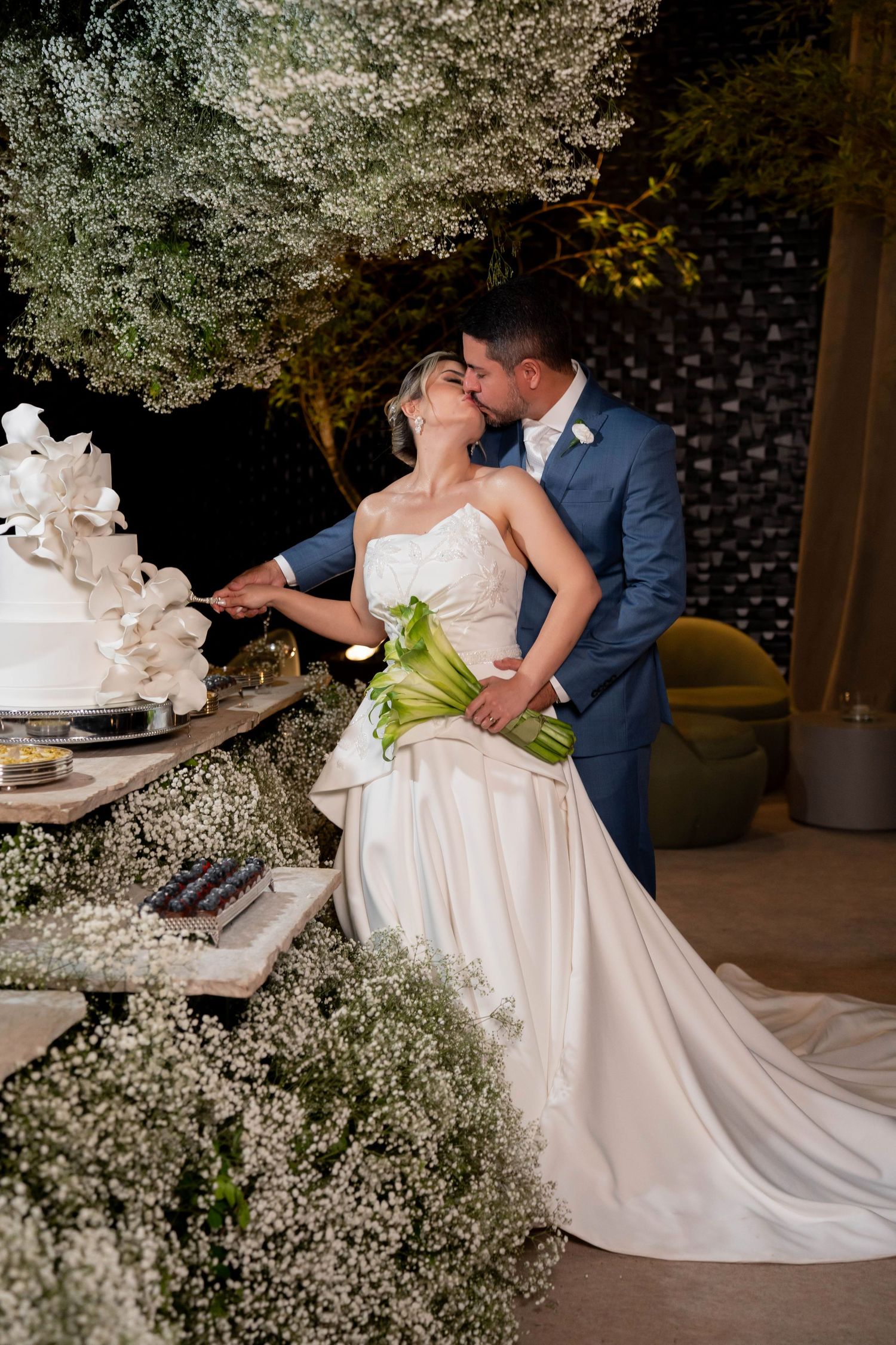 Bride and groom share a kiss while cutting their white floral wedding cake, surrounded by baby's breath flowers.