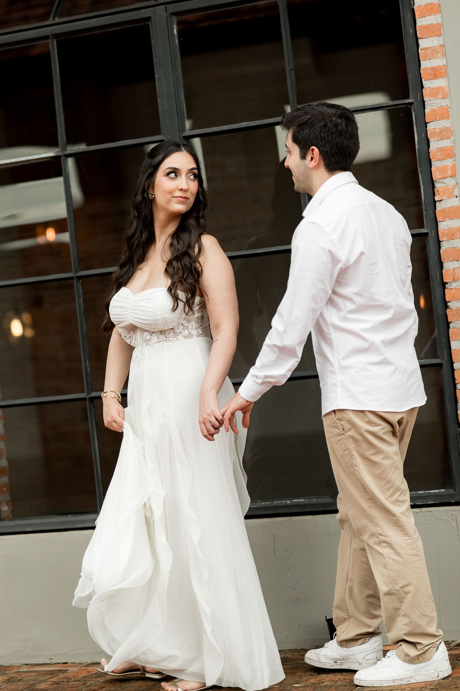 Bride in white gown holds hands with groom in white shirt and khakis outside brick building.