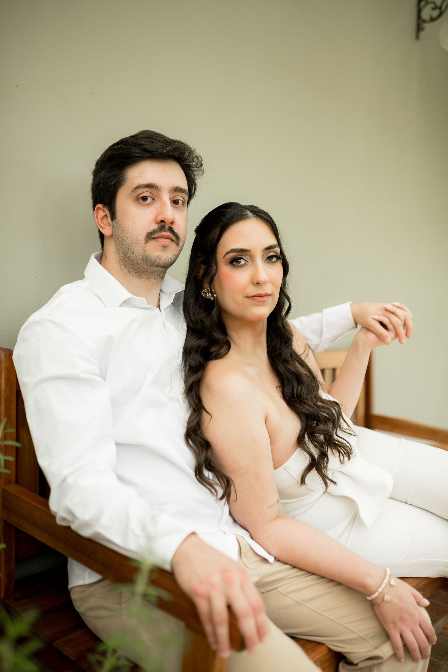 Elegant couple in white outfits sitting on a wooden bench, woman with long wavy hair, man with mustache.