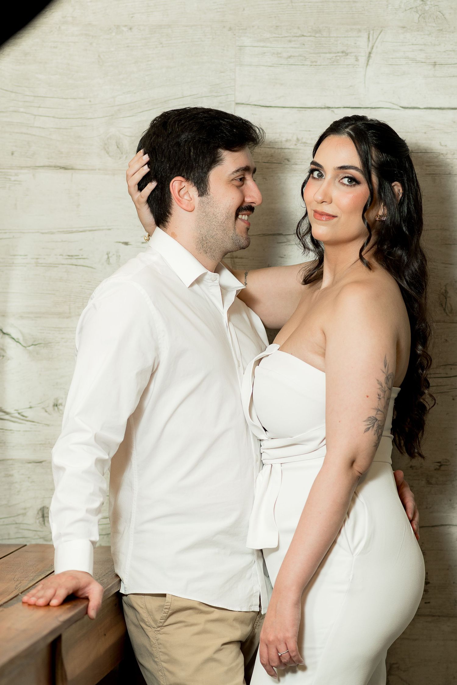 A couple in white outfits sharing a romantic moment, the woman with a tattoo embracing the smiling man against a rustic wall.