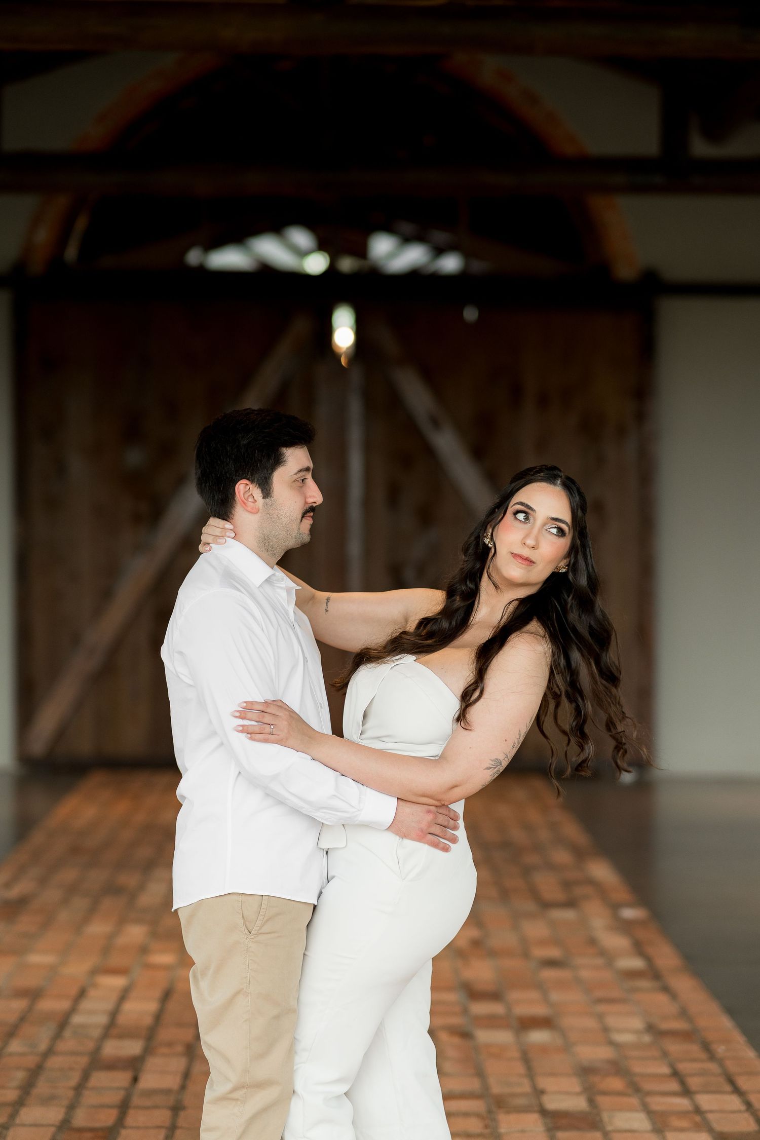 Couple embracing in a rustic barn venue, woman in white dress with long dark hair, man in white shirt and khaki pants.