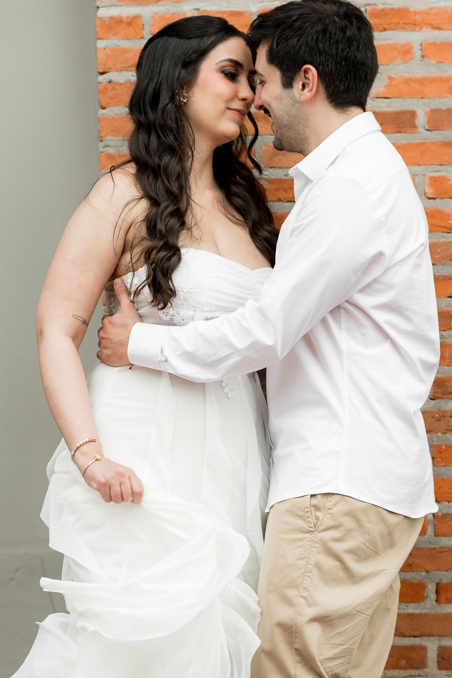 A couple in wedding attire share a romantic moment against a brick wall, the bride in white gown, groom in white shirt.