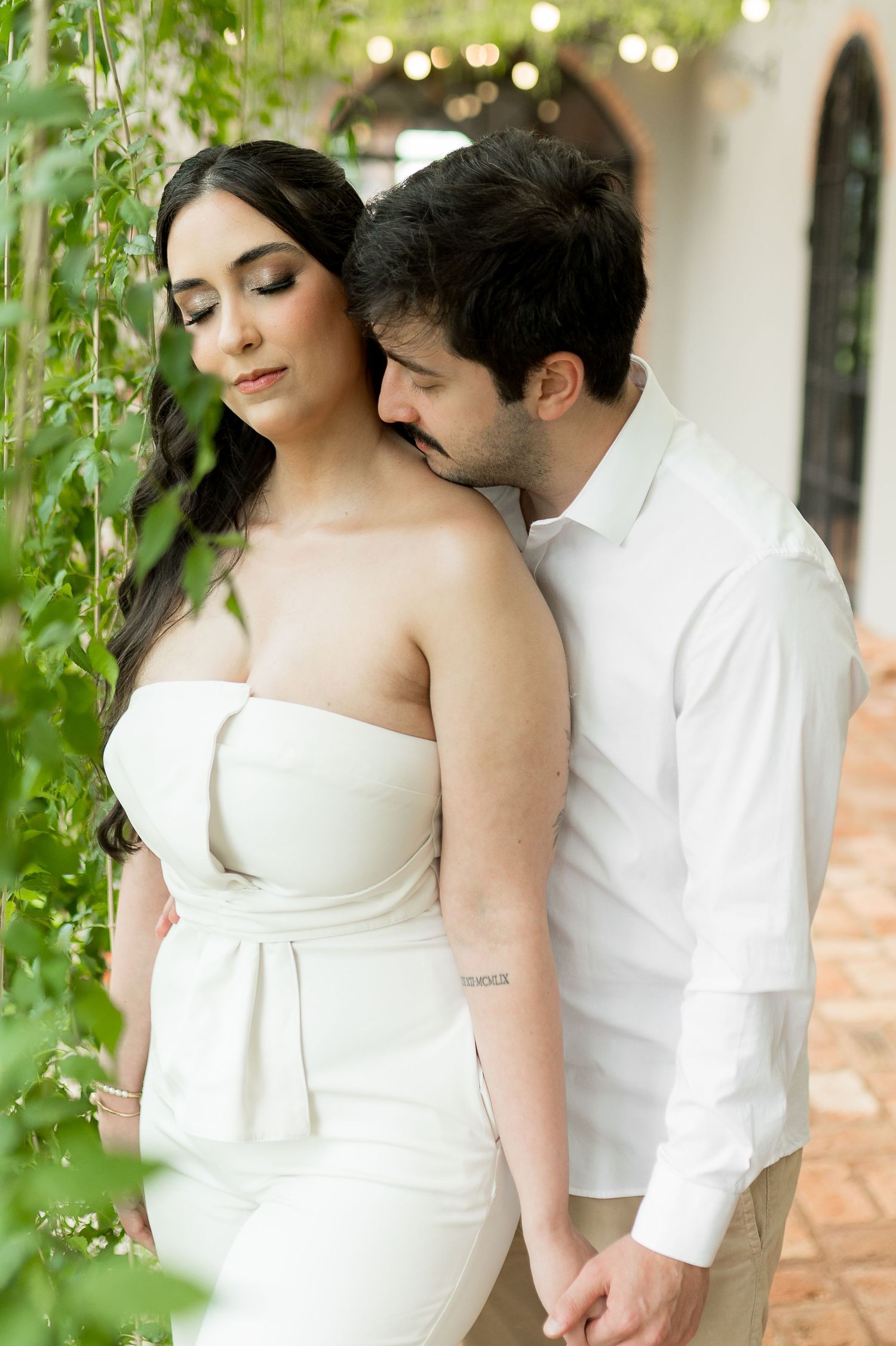 Romantic couple in white outfits embracing among green vines, woman in strapless dress, man in white shirt, soft bokeh lights.