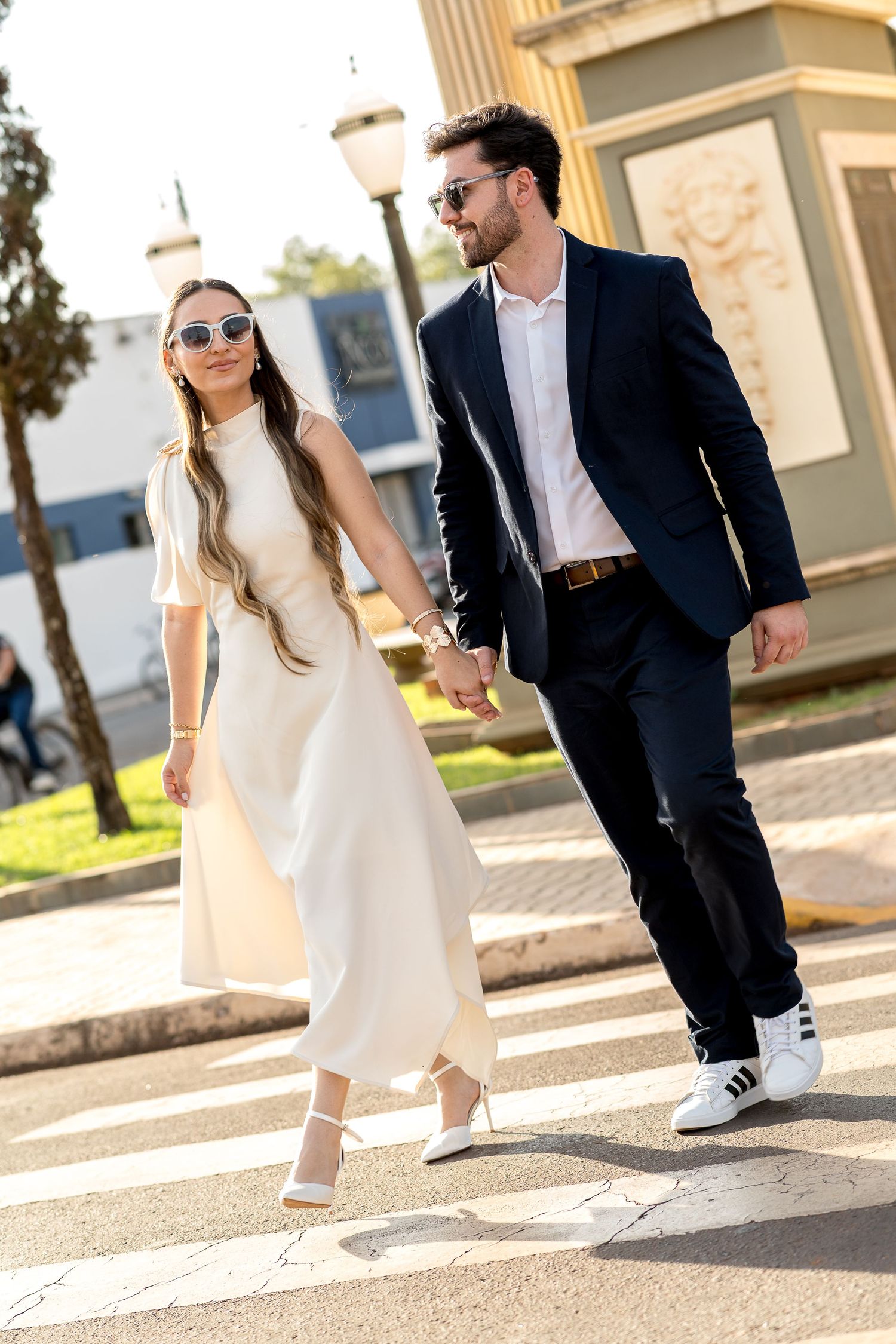 Elegant couple holding hands crossing a street, woman in white dress and man in navy suit, both wearing sunglasses.
