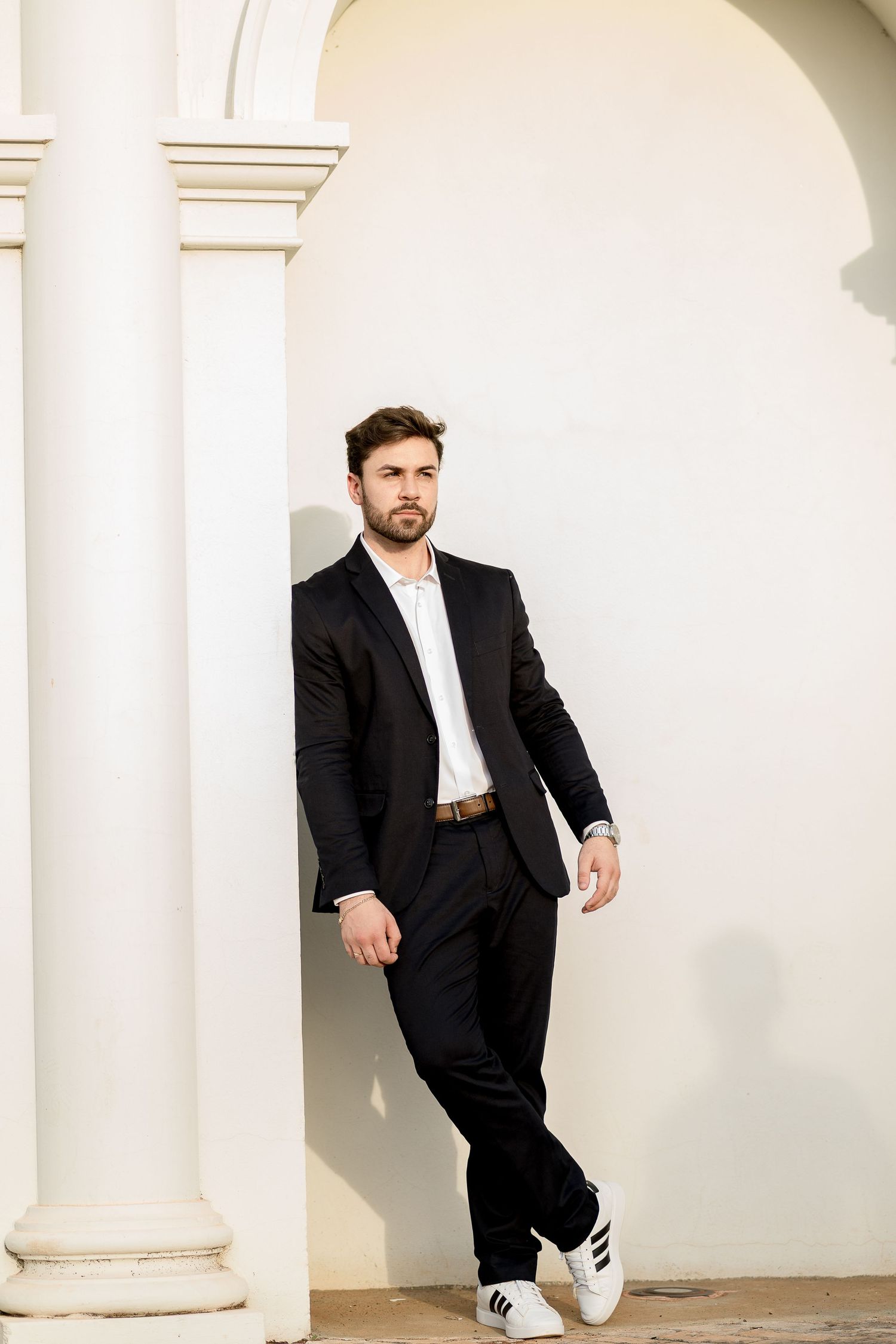 A stylish man in a black suit and white sneakers leans against a white wall with elegant columns.
