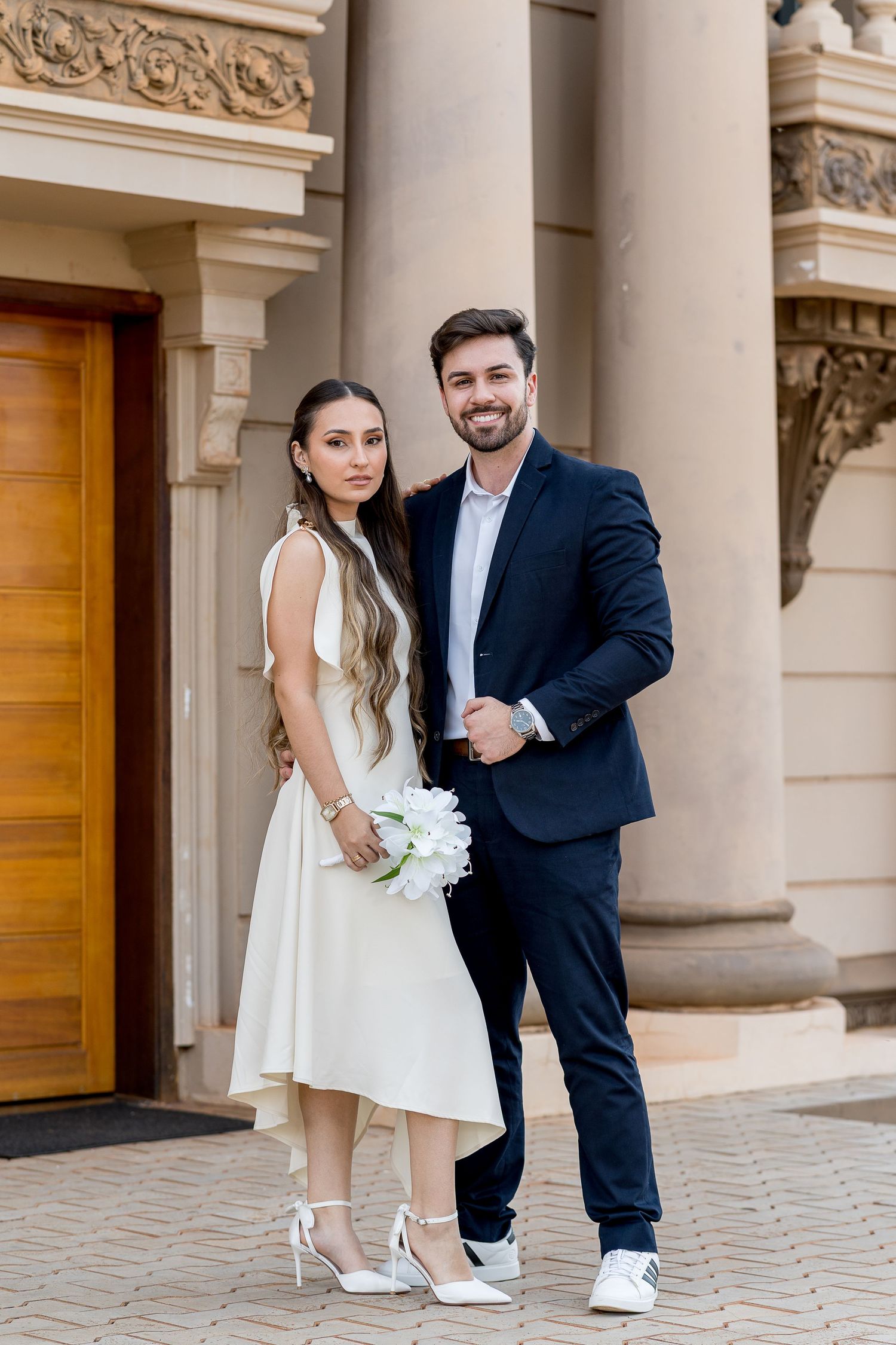 Smiling newlyweds stand before ornate building columns, bride in white midi dress with bouquet, groom in dark navy suit.