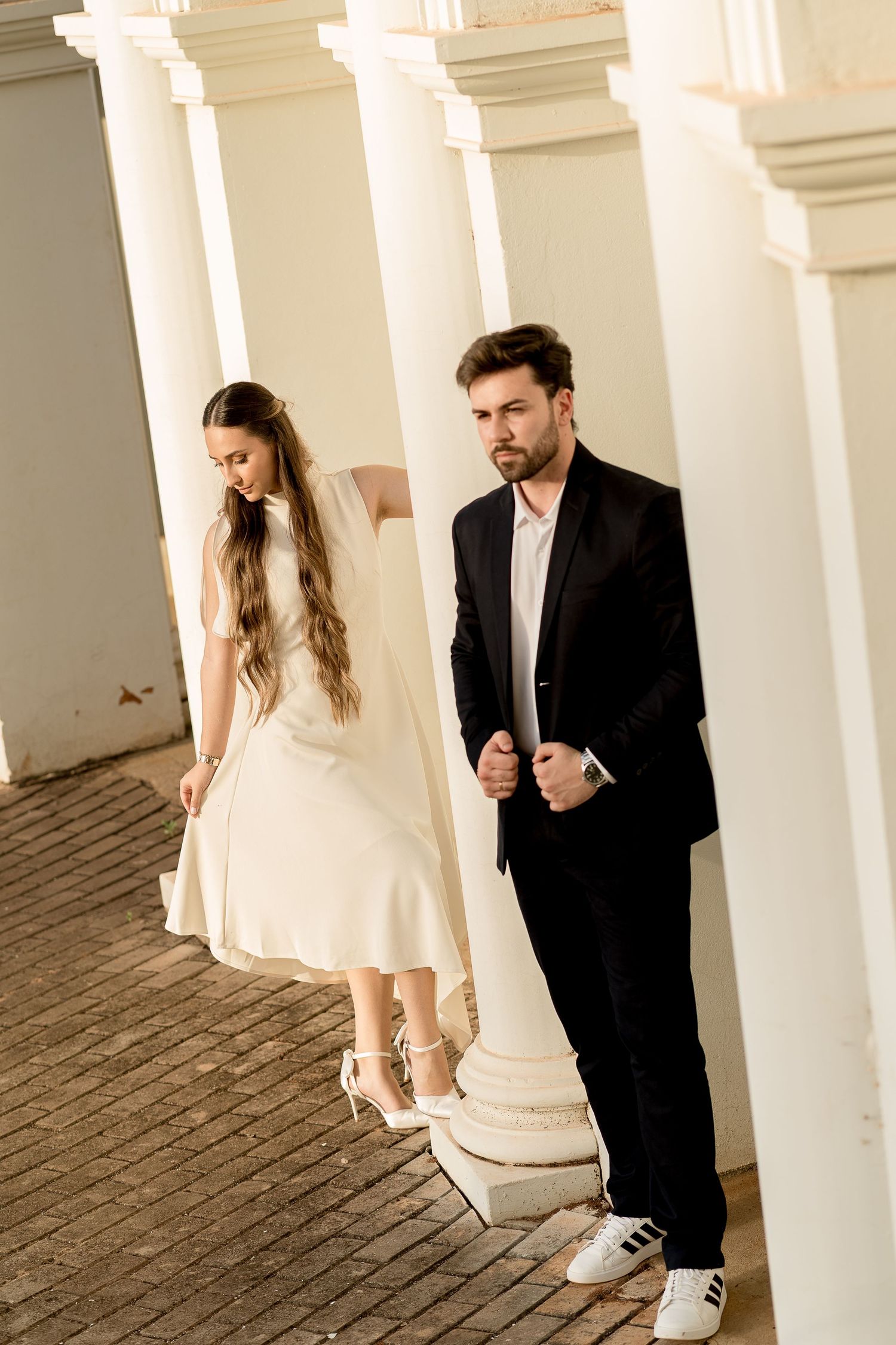 Elegant couple in wedding attire posing between white columns, groom in black suit, bride in white dress.