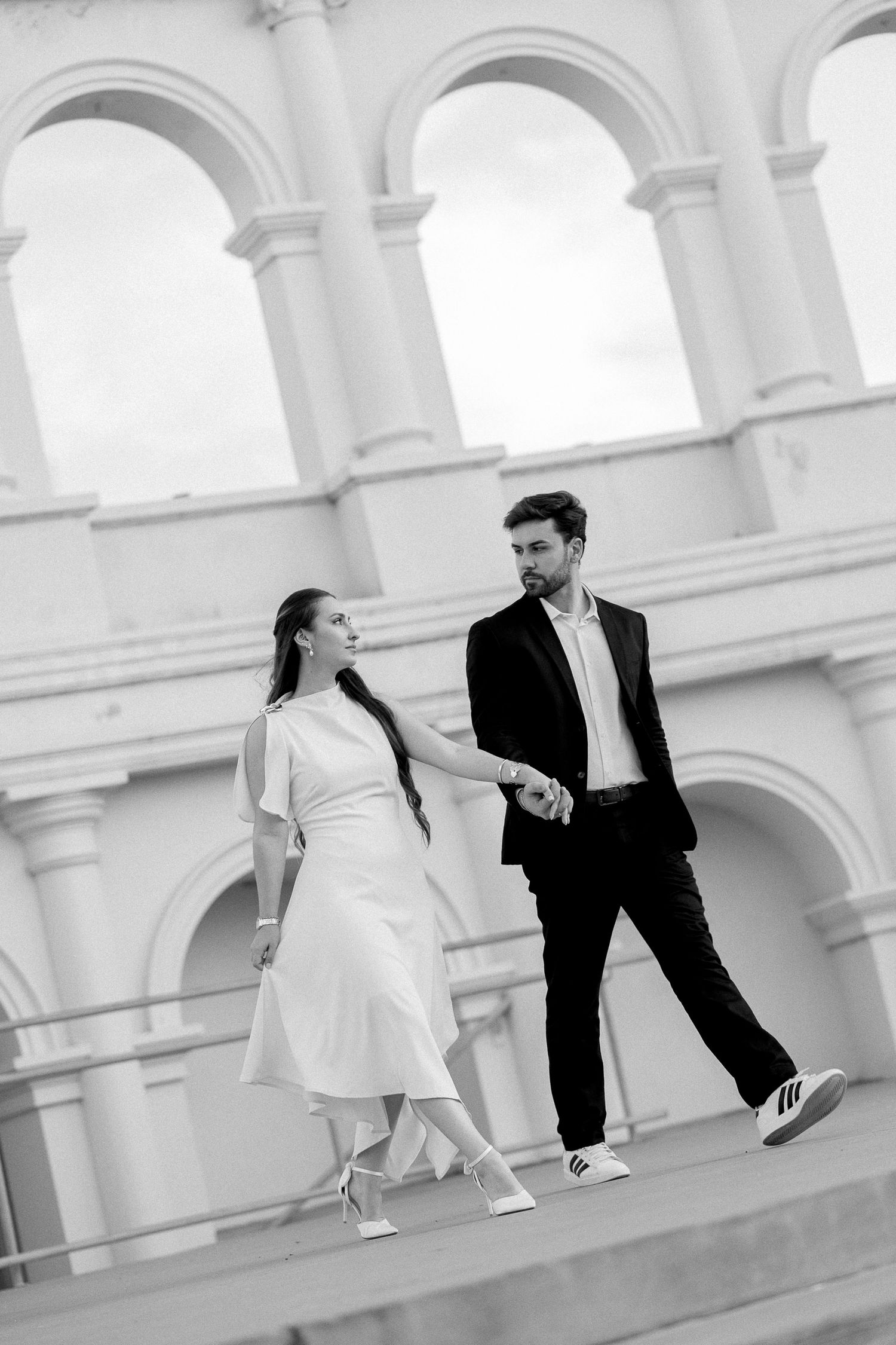 Black and white photo of an elegant couple holding hands walking in front of a classical arched building.