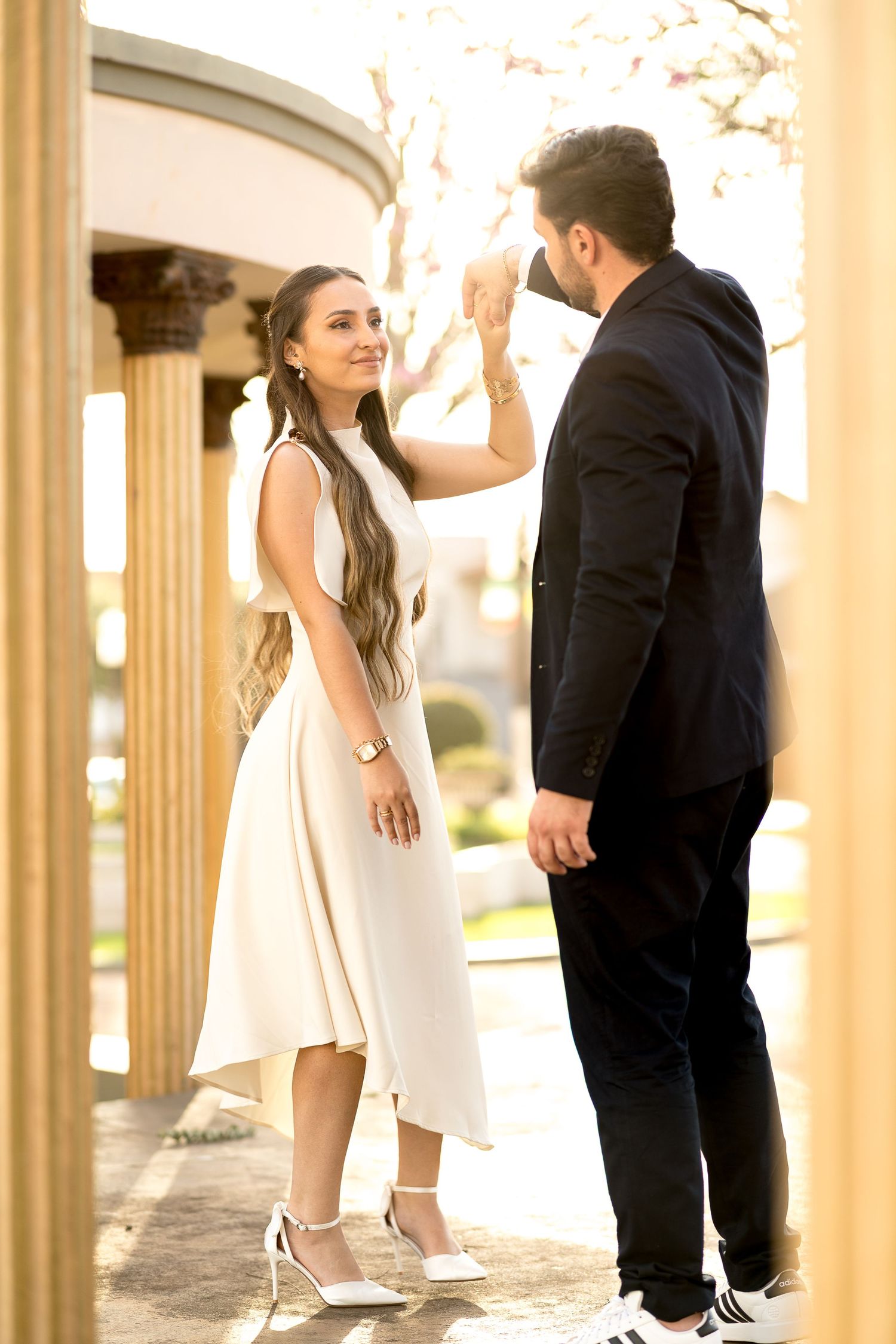 Elegant couple dancing outdoors, woman in white midi dress and heels, man in dark suit, surrounded by classical columns.