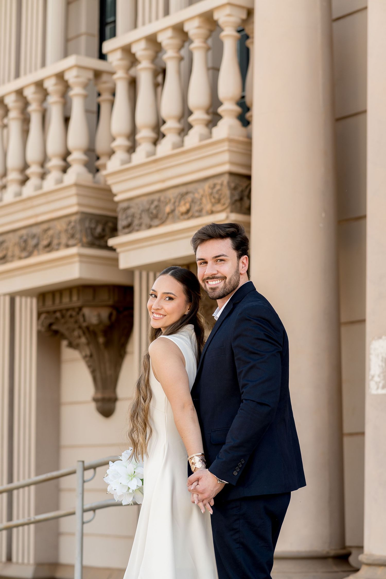 A smiling bride and groom hold hands in front of an elegant beige building with ornate columns and balustrades.