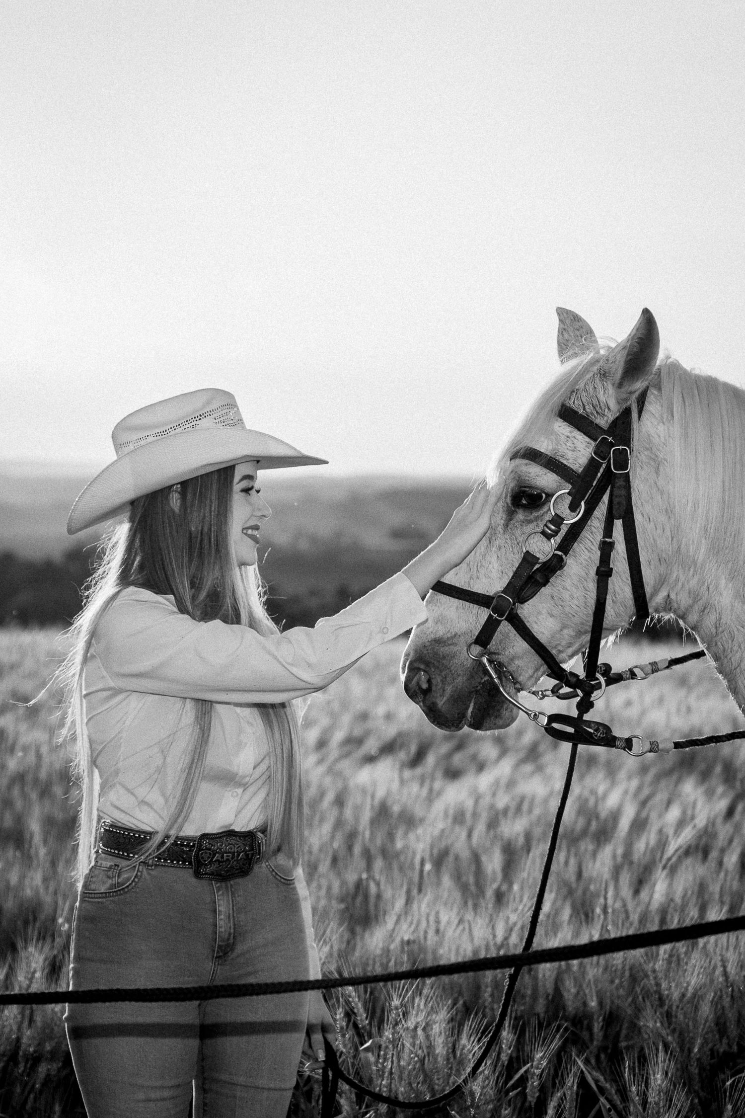 Black and white photo of a cowgirl in a hat petting a white horse in a field at sunset.