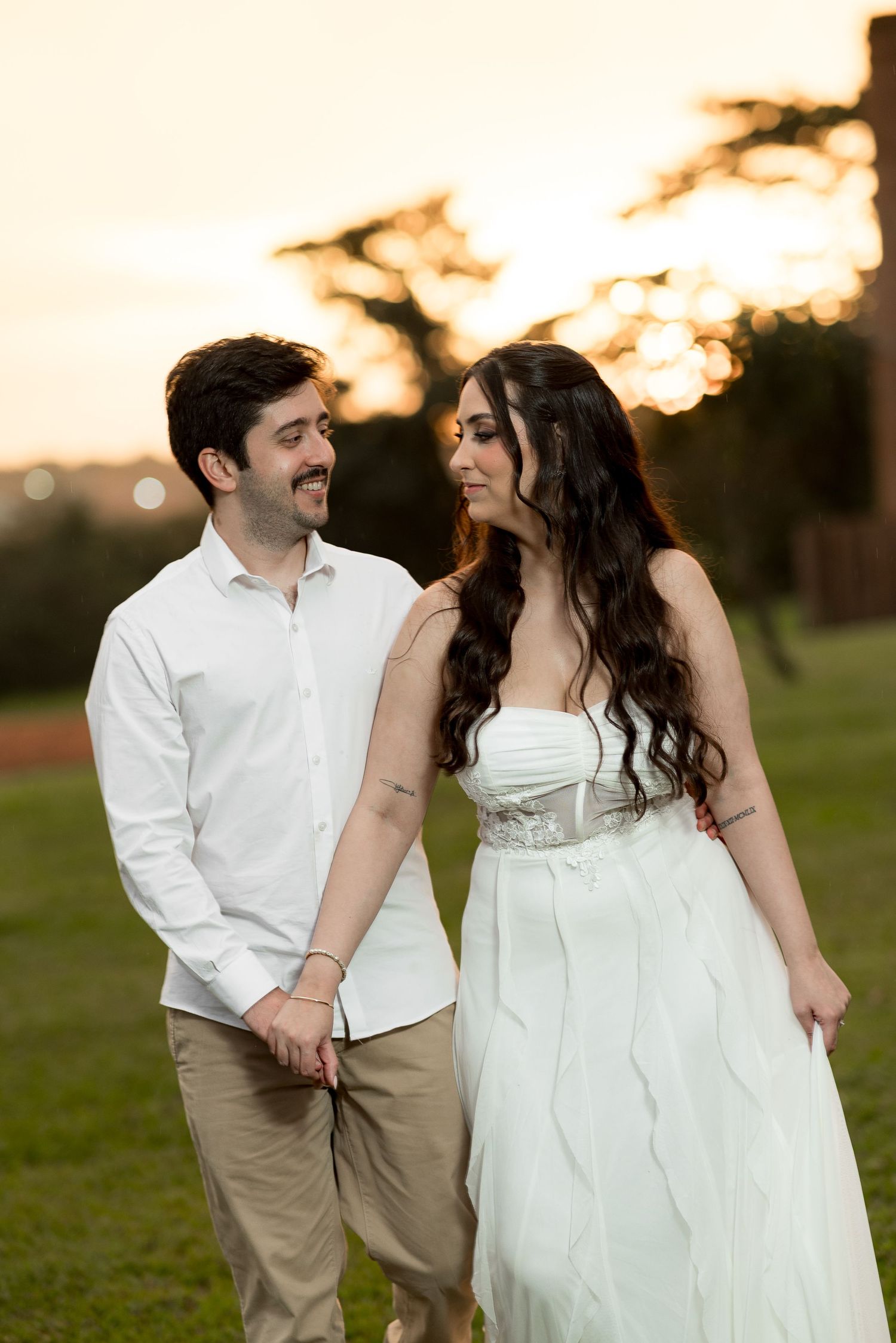 Couple in wedding attire holding hands at sunset, bride in white gown, groom in white shirt and khakis.