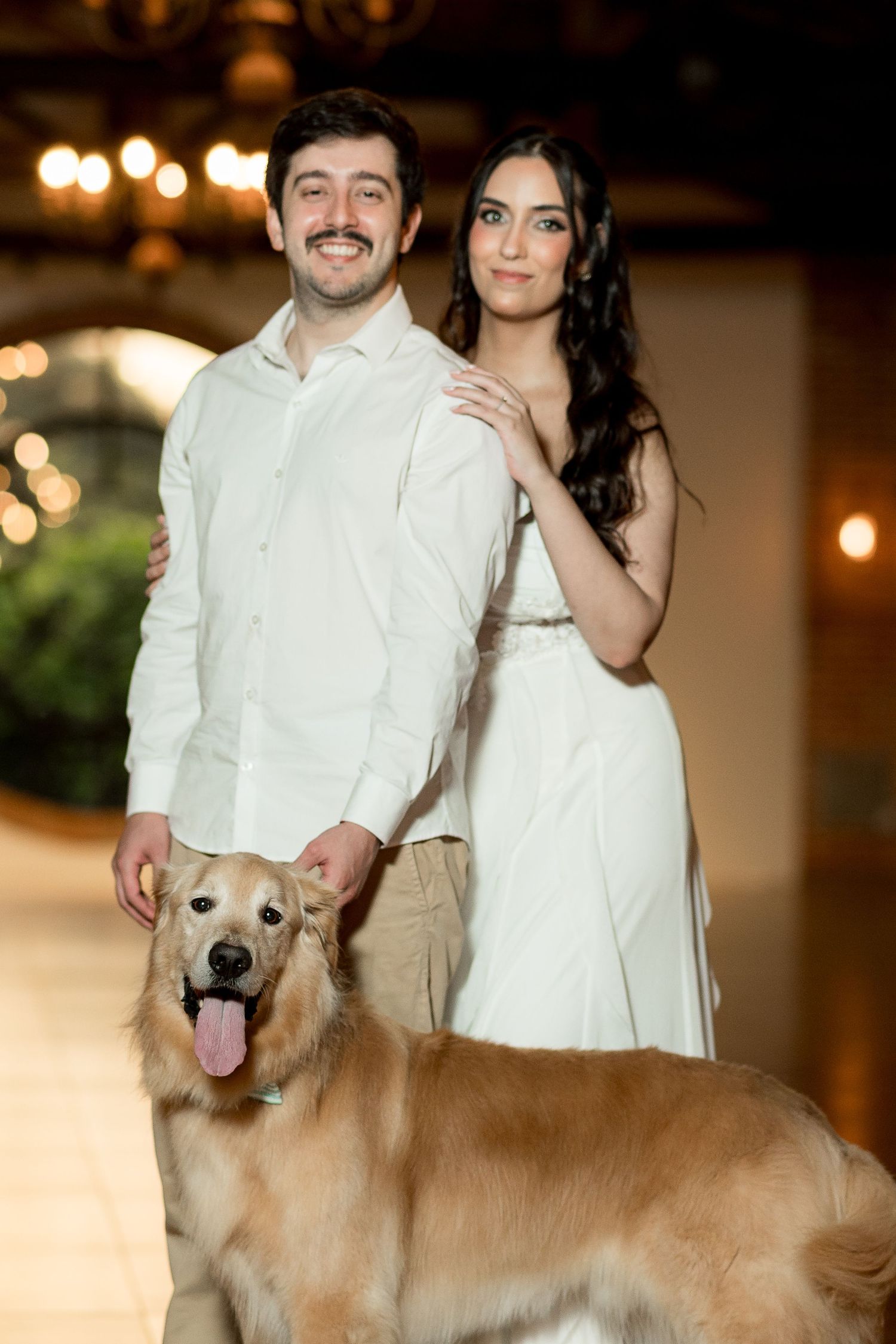 Couple in white attire posing with a golden retriever dog in an elegantly lit indoor venue.
