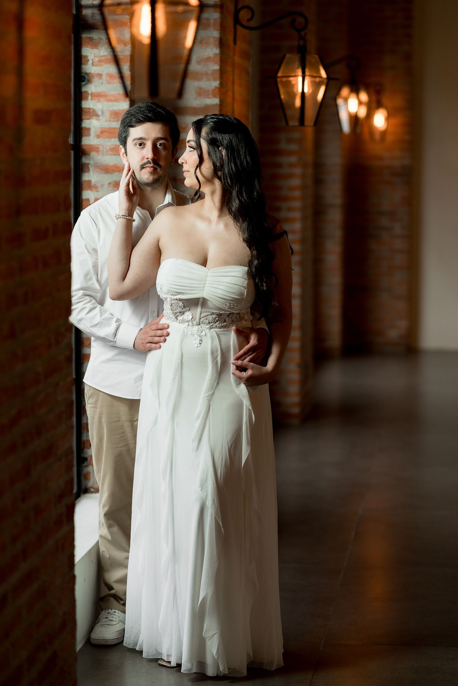 Romantic couple in wedding attire embracing near brick wall with vintage lanterns, bride in white strapless gown.