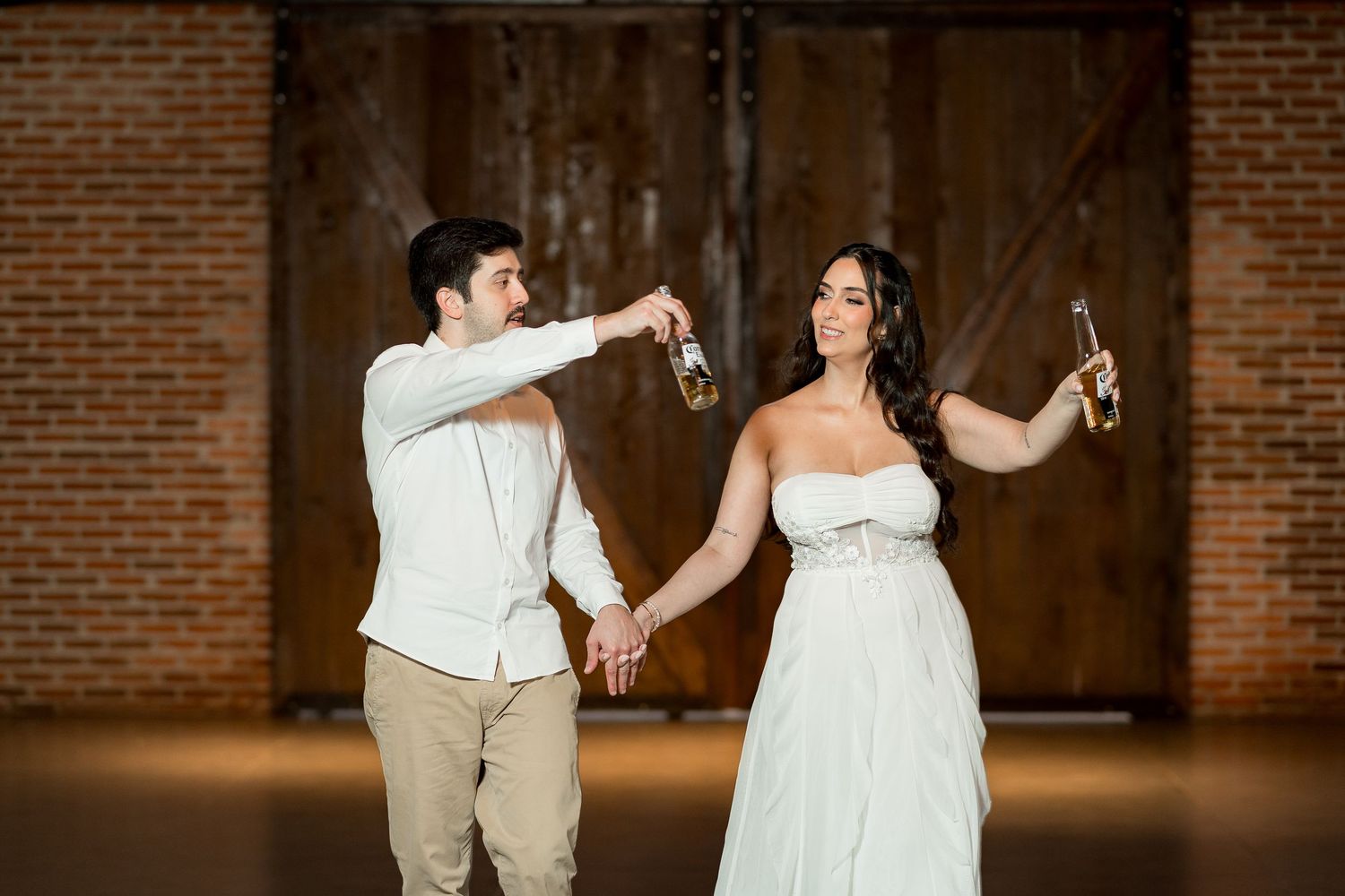 Bride and groom toasting with beer bottles in a rustic barn wedding venue with exposed brick walls.