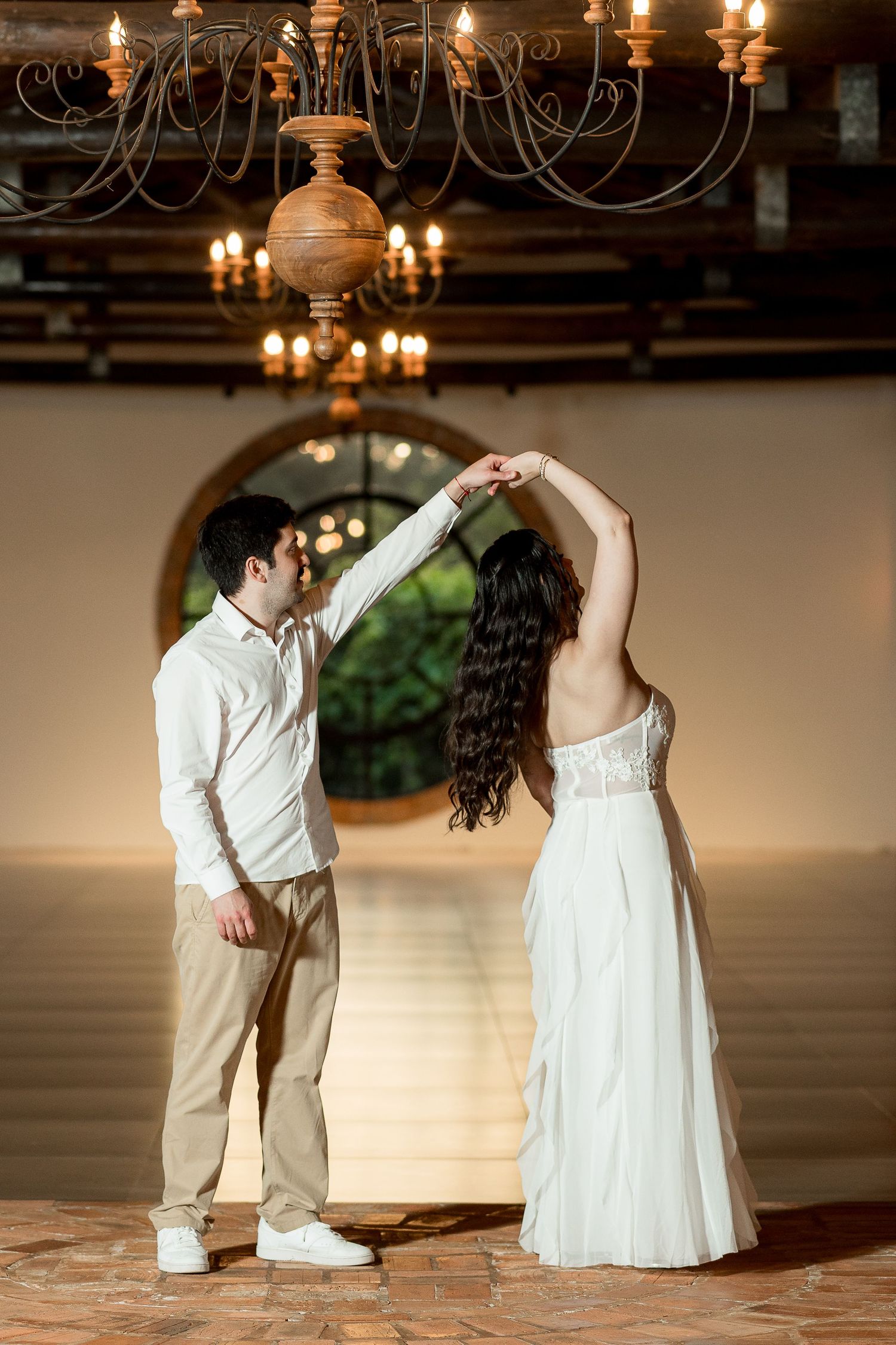 Groom spinning bride in white gown under ornate chandelier in romantic wedding venue dance floor.