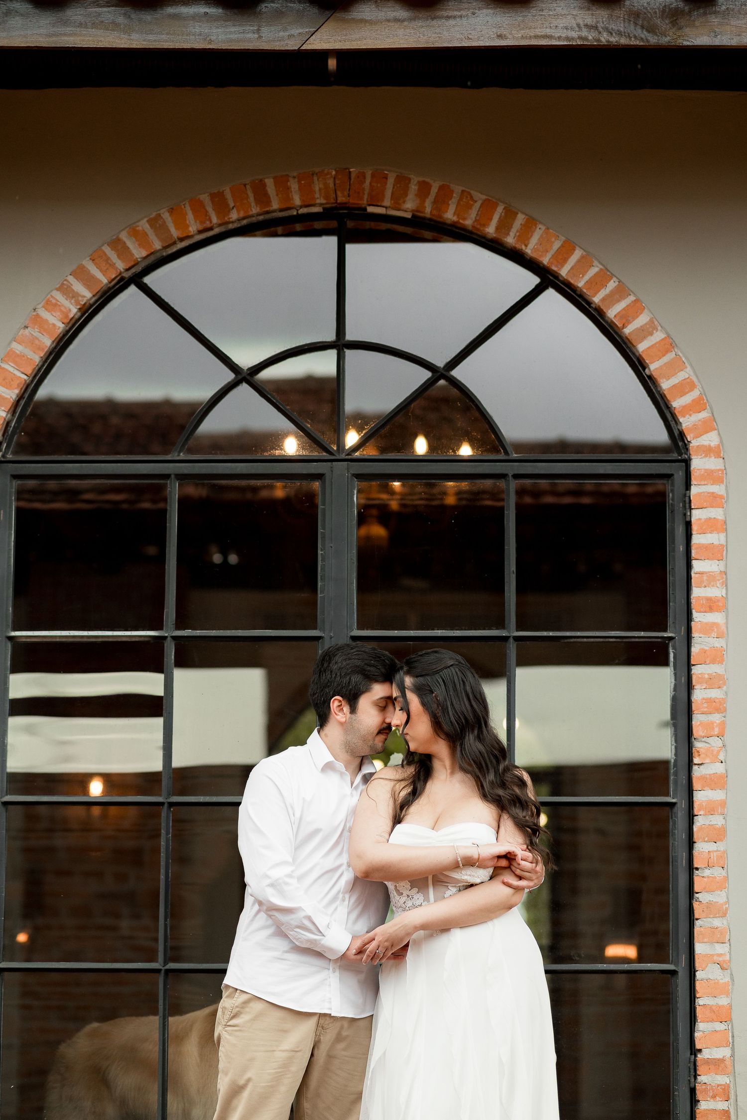 Couple sharing a romantic moment in front of an arched brick window, man in white shirt, woman in white strapless dress.