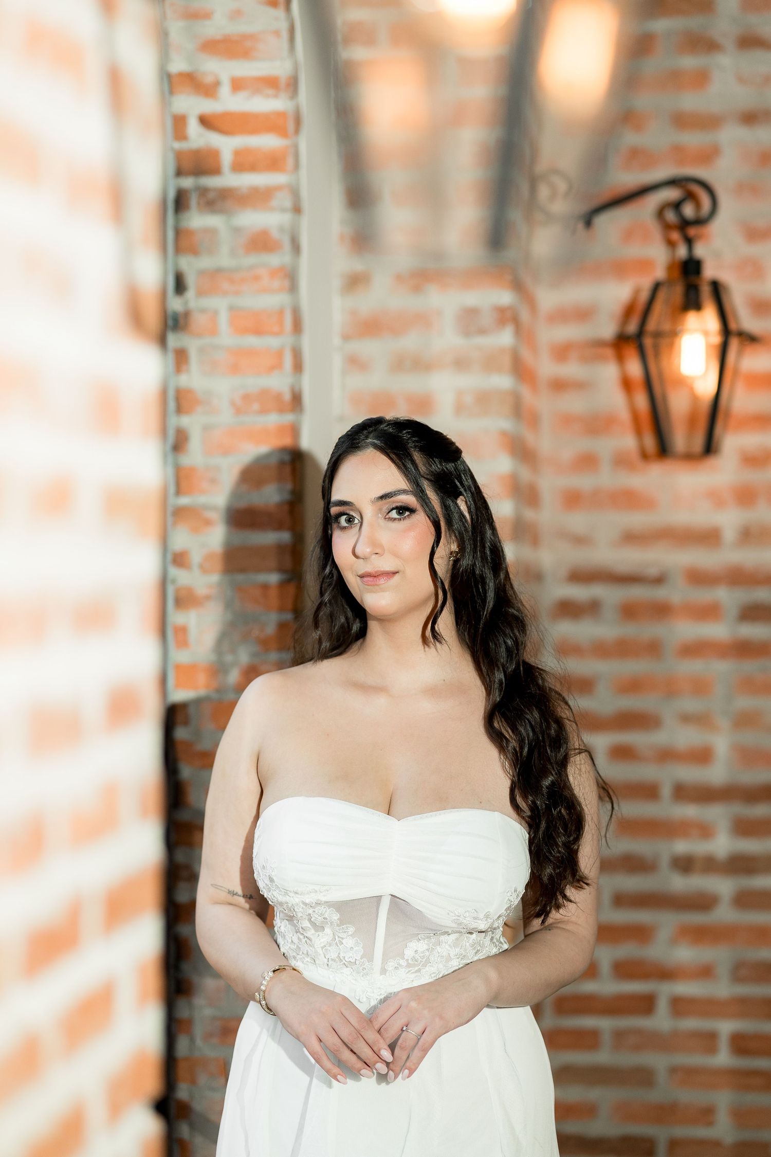 Bride in strapless white wedding gown poses gracefully against rustic brick wall with vintage lantern lighting.