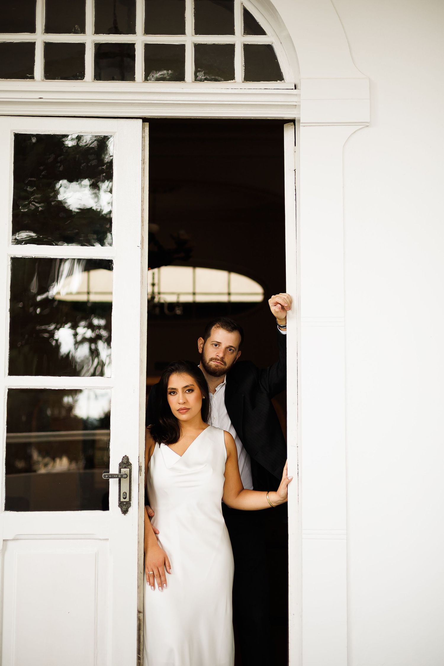 Bride in white gown and groom in black suit posing elegantly in a white doorway for their wedding portrait.