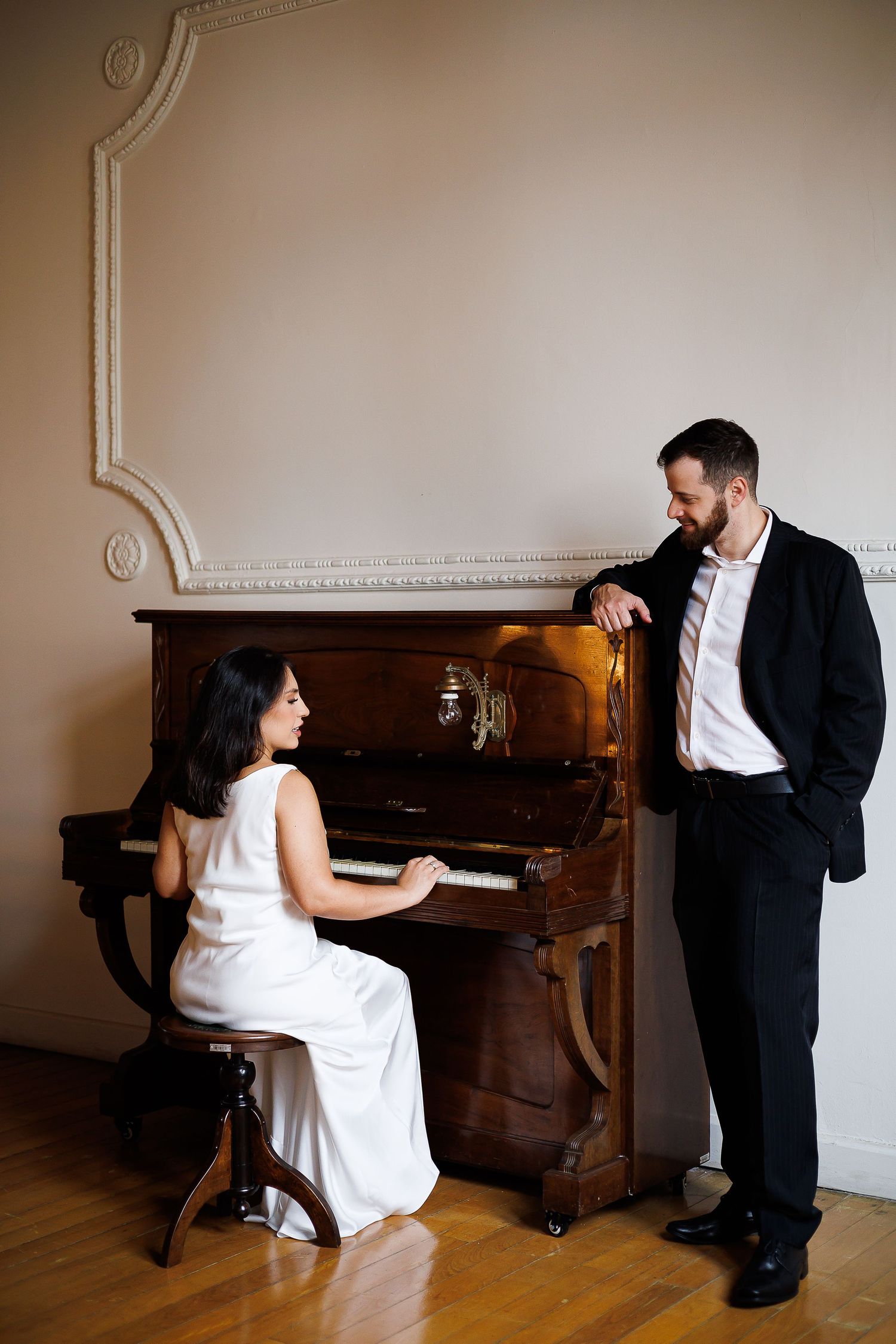 A woman in a white dress plays an upright piano while a man in a black suit leans against it smiling.