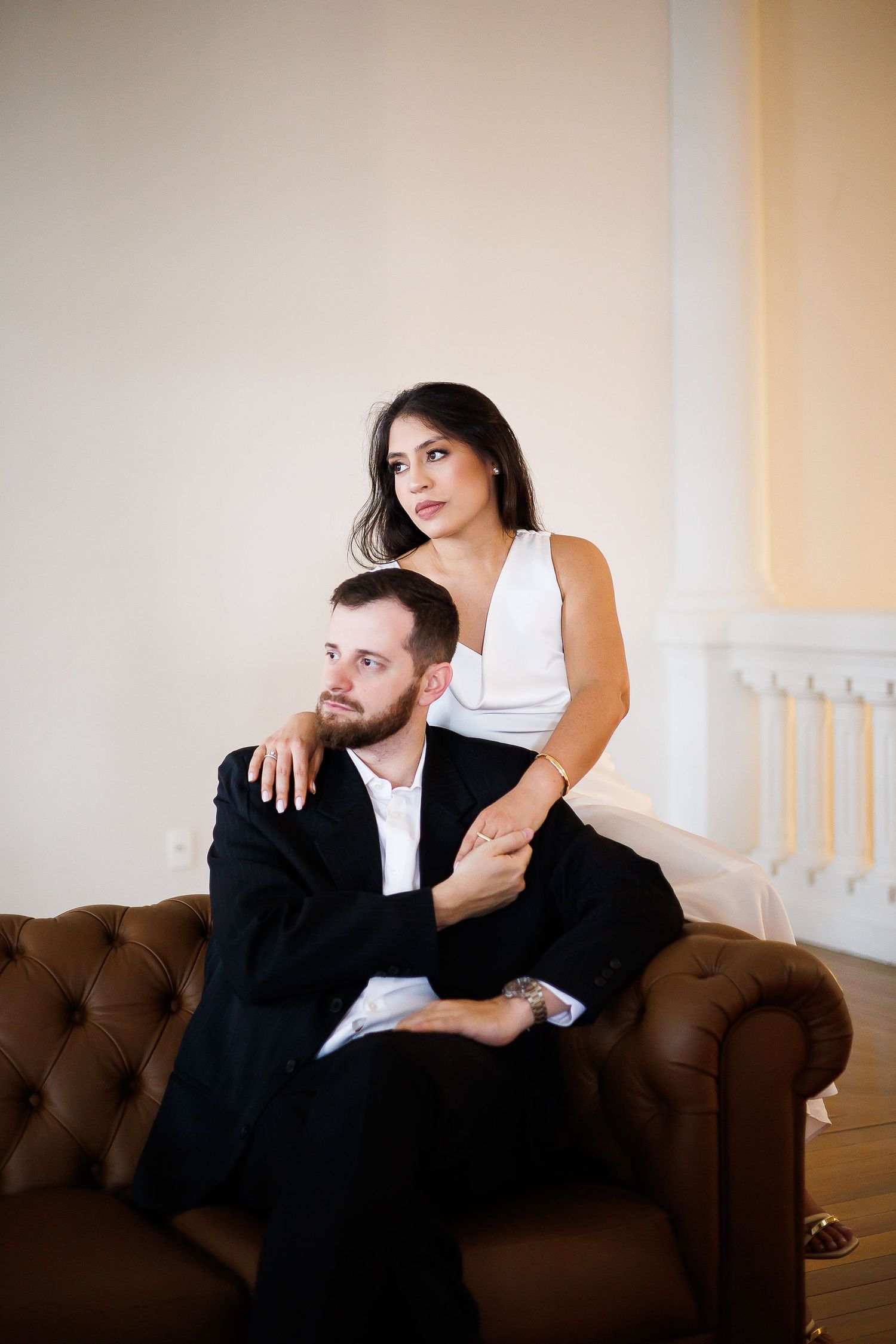 Elegant couple posing in a luxurious interior, man in black suit seated on leather sofa, woman in white dress embracing him.