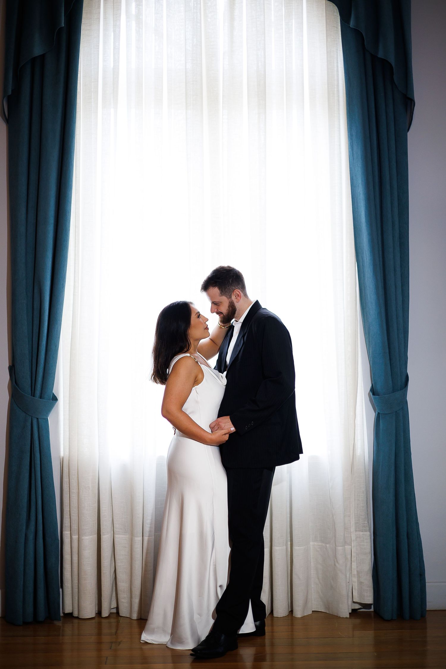 Elegant couple in formal wear embracing before a large window with blue curtains in a romantic indoor setting.