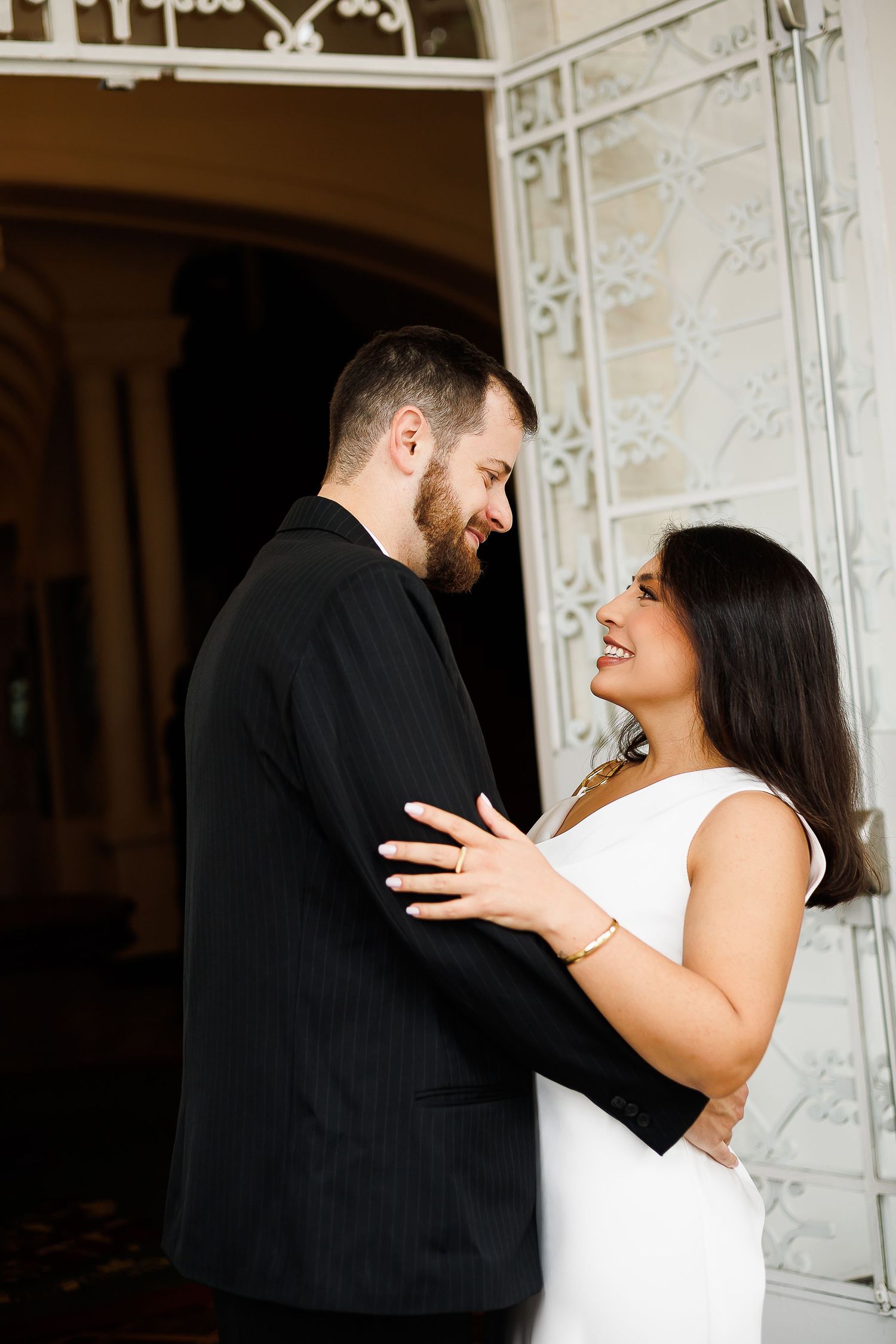 Couple in elegant attire embracing and smiling at each other near ornate white architecture.