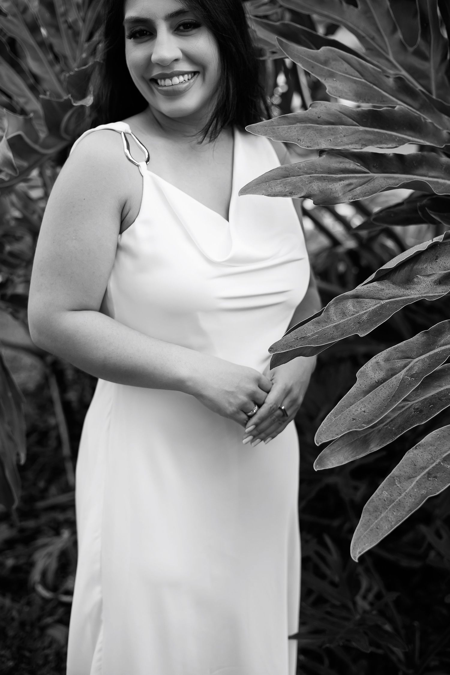 Black and white photo of a smiling woman in a white gown posing among tropical leaves.