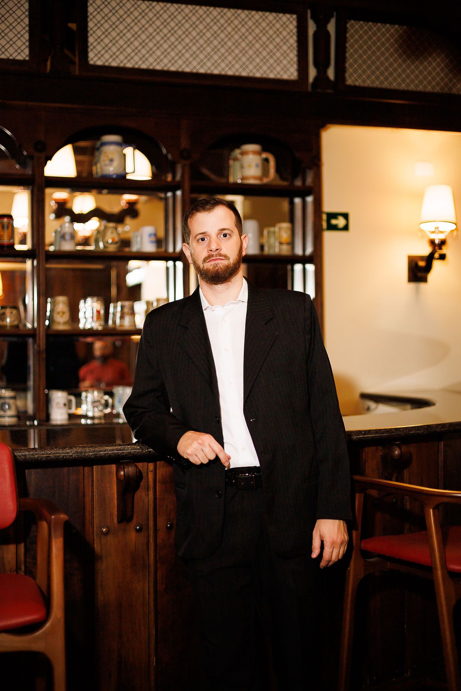 Bearded man in black suit standing at a wooden bar with shelves displaying mugs and warm lighting.