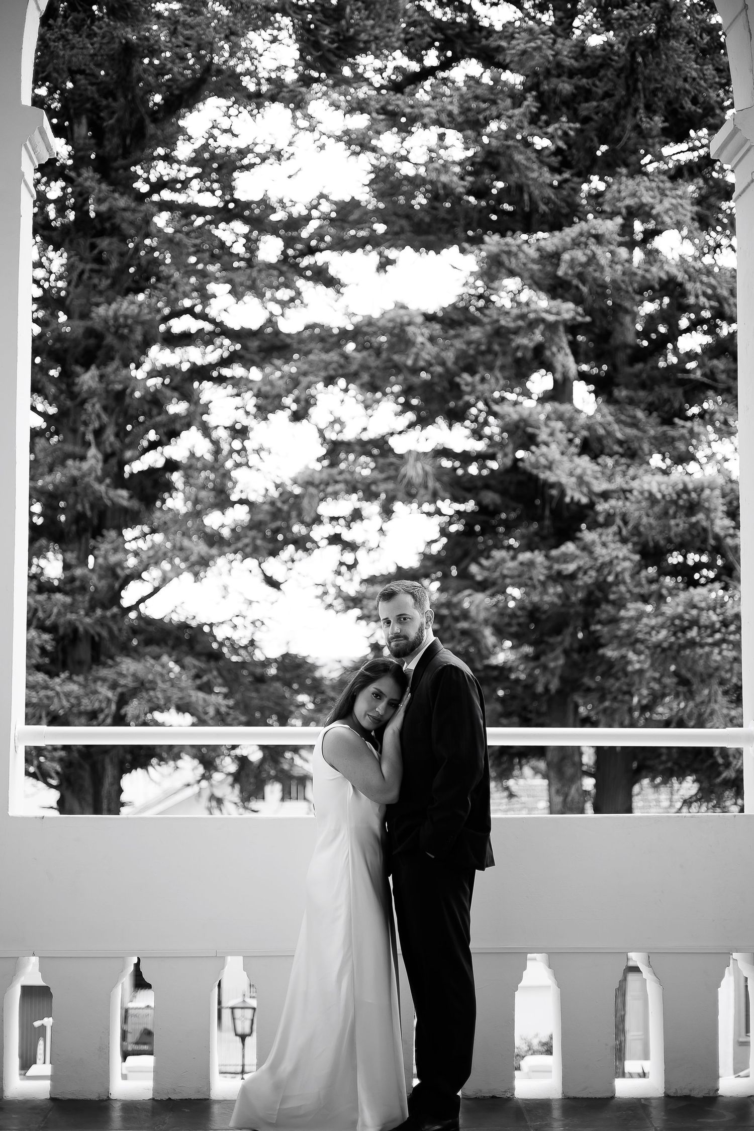 Black and white wedding photo of bride and groom embracing on a balcony with lush trees in the background.