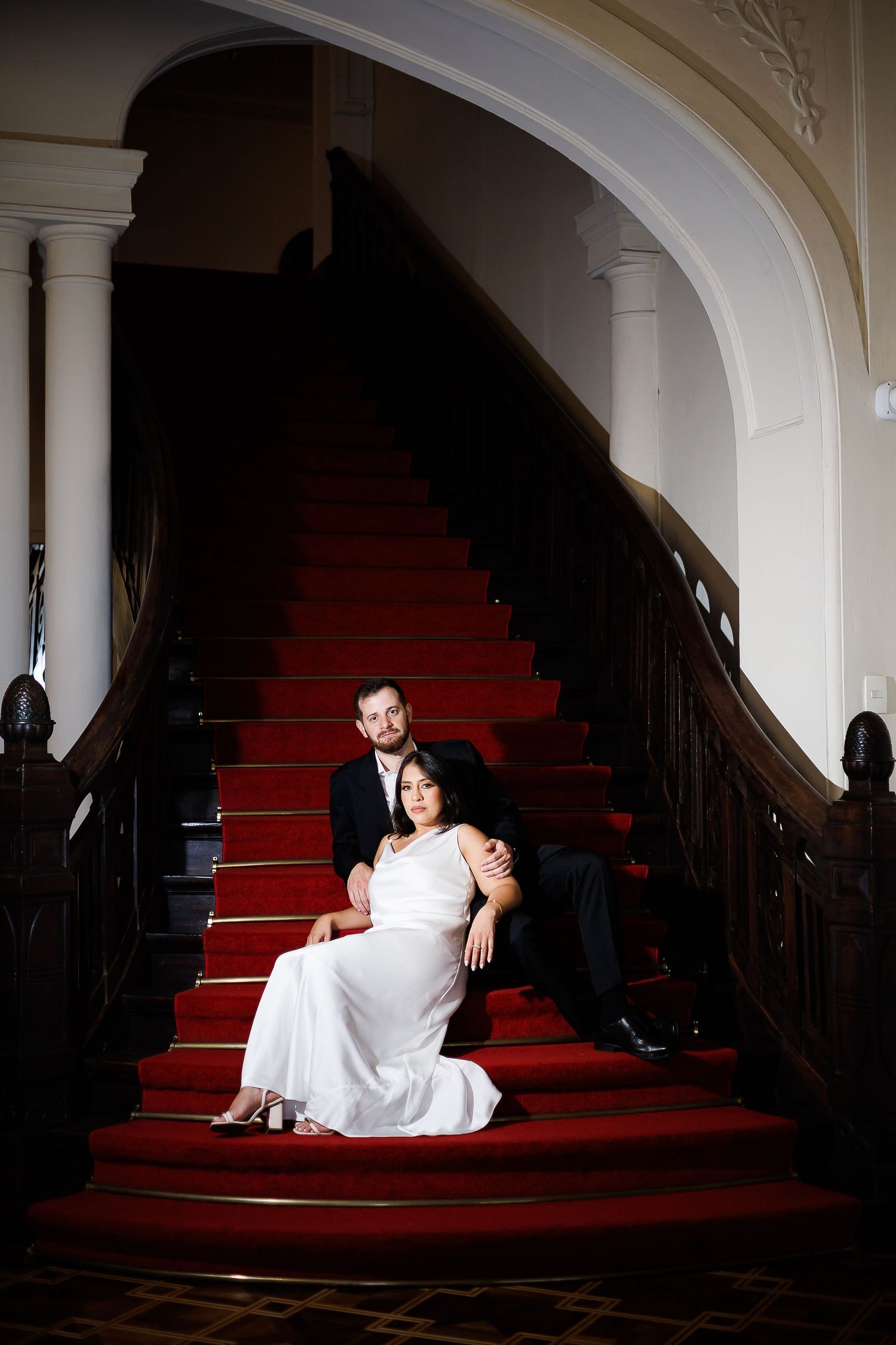Elegant couple in wedding attire posing on red carpeted grand staircase in a classic venue.