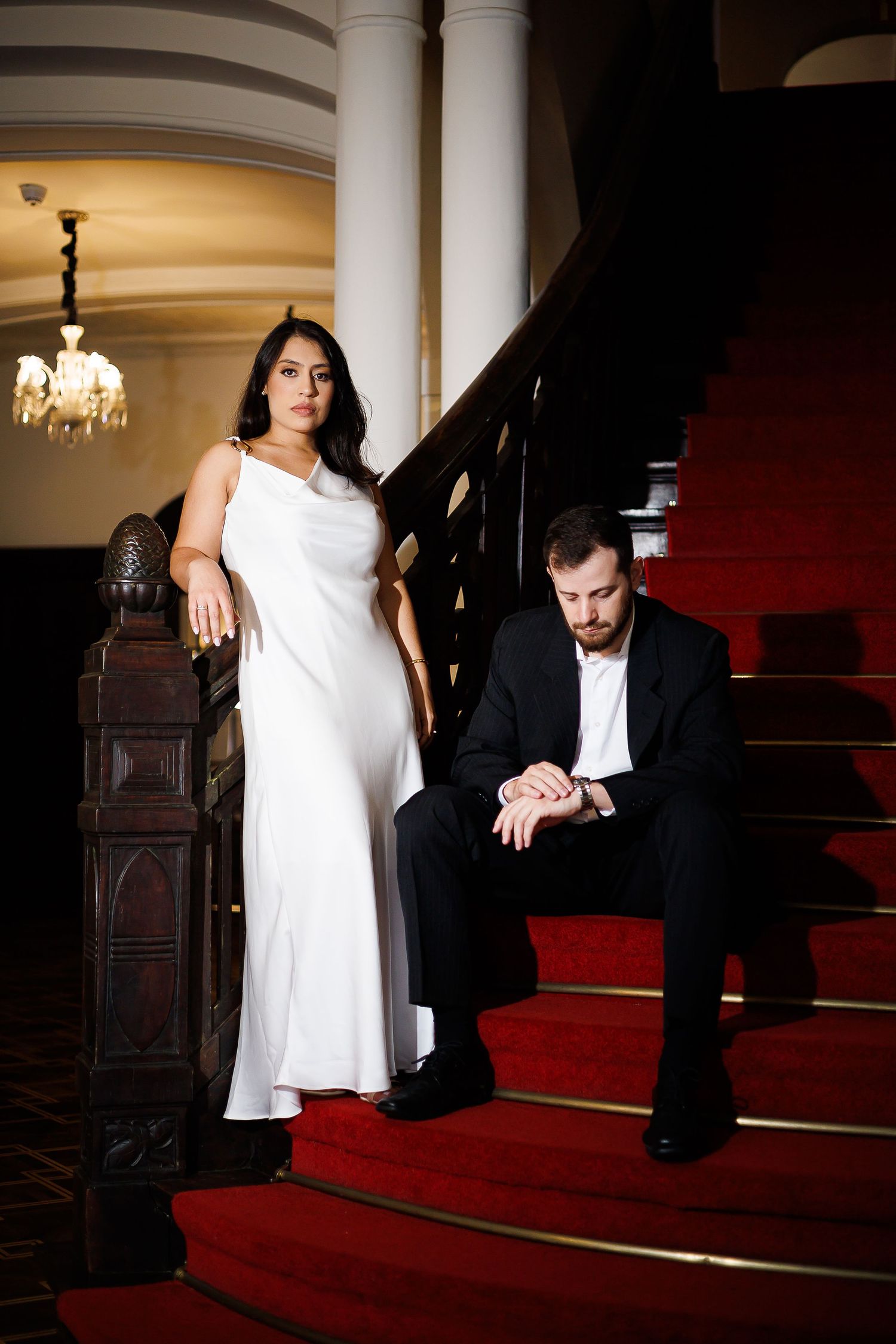 Elegant couple on red carpeted stairs, woman in white gown, man in black suit checking watch in grand hall.