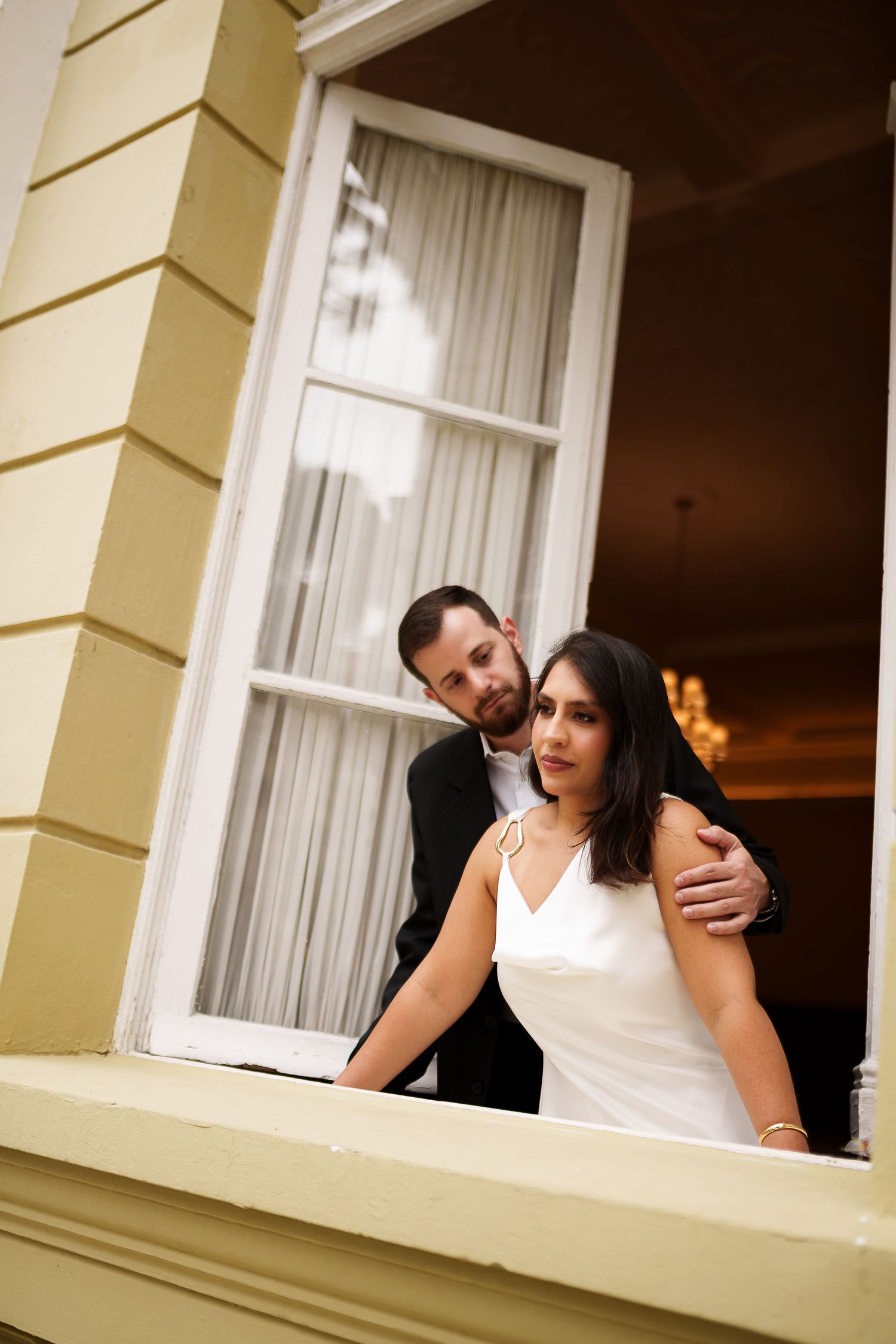 A bride and groom pose romantically on a balcony, she in a white gown and he in a dark suit, at an elegant venue.