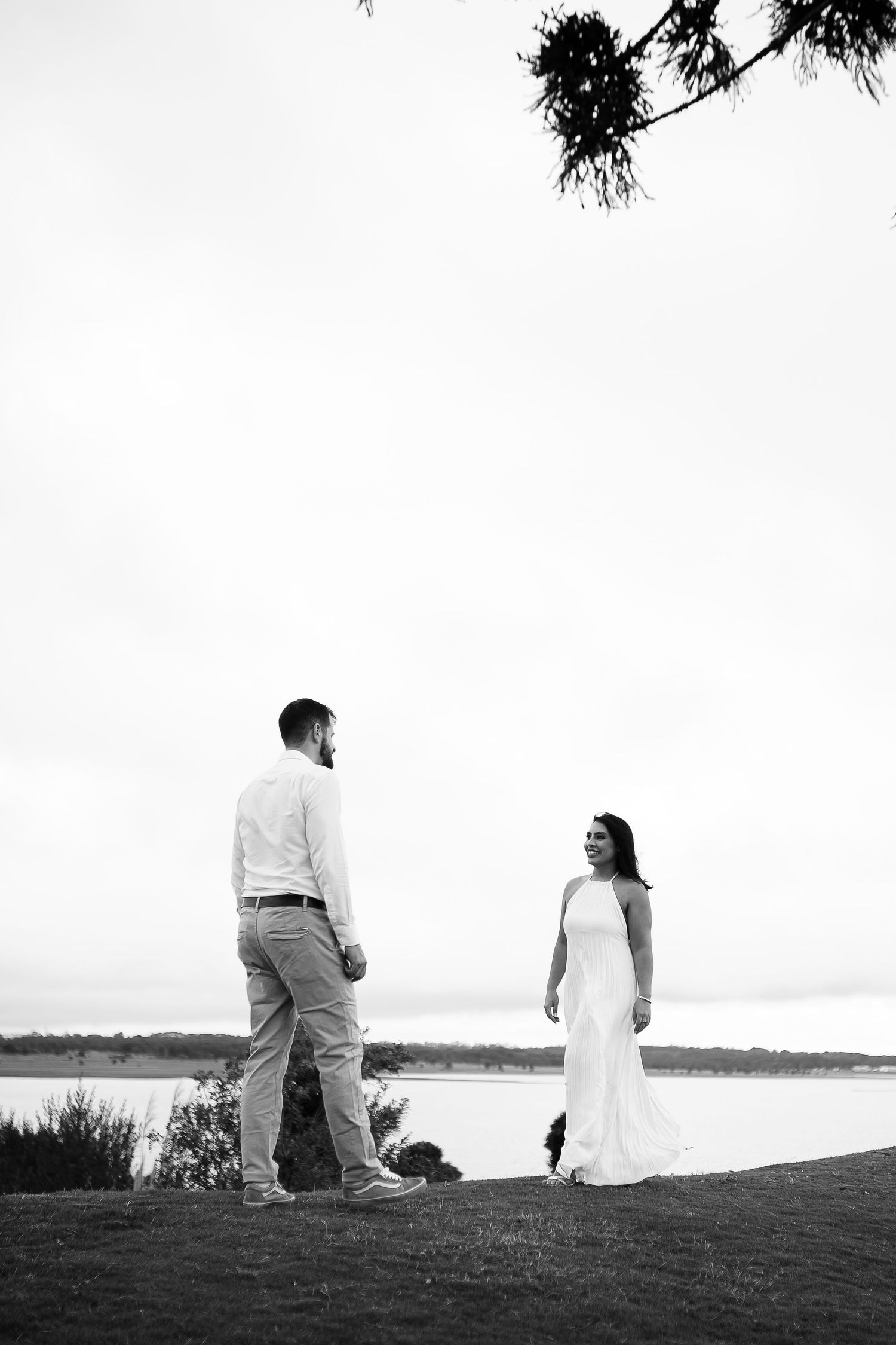 Black and white photo of a bride in a white gown and groom in casual attire standing by a serene lakeside.