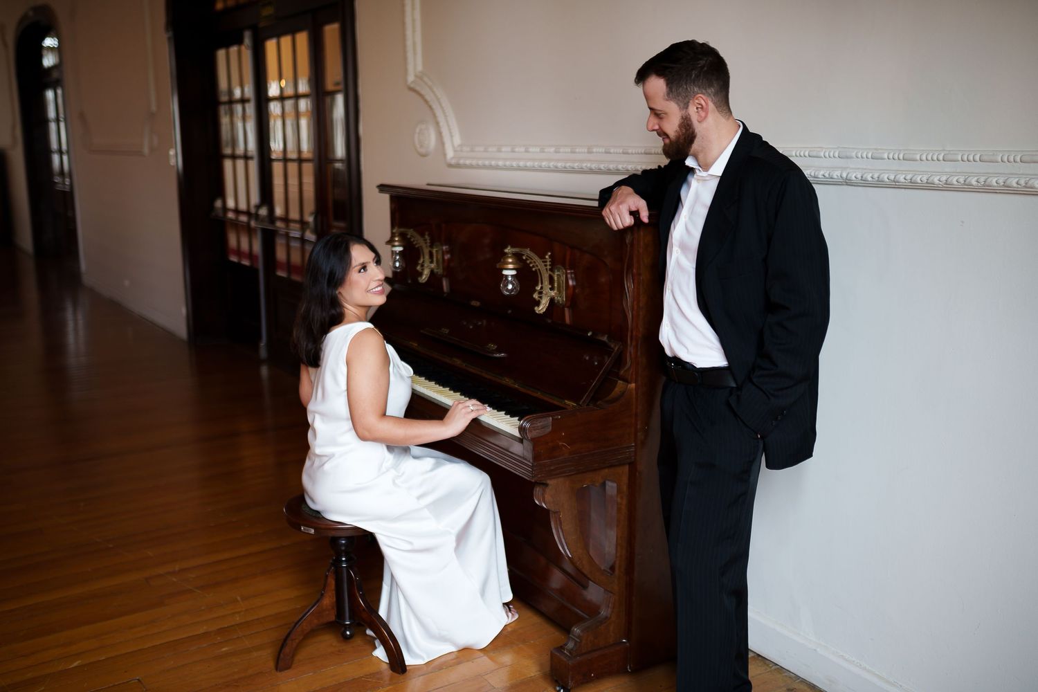 Bride in white dress playing upright piano while groom in black tuxedo stands beside her in elegant room.
