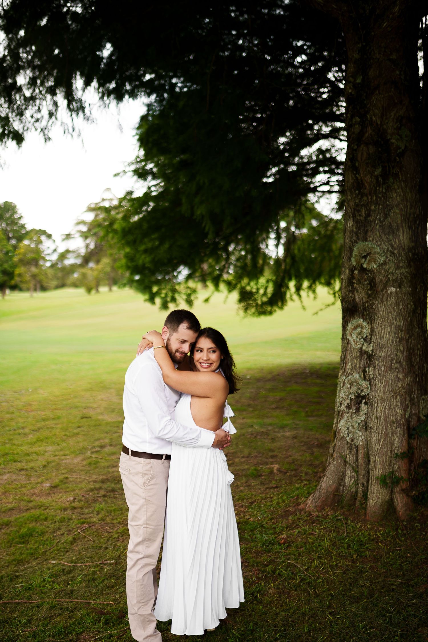 Couple embracing near a large tree on a lush green lawn, woman in white dress smiling at camera.