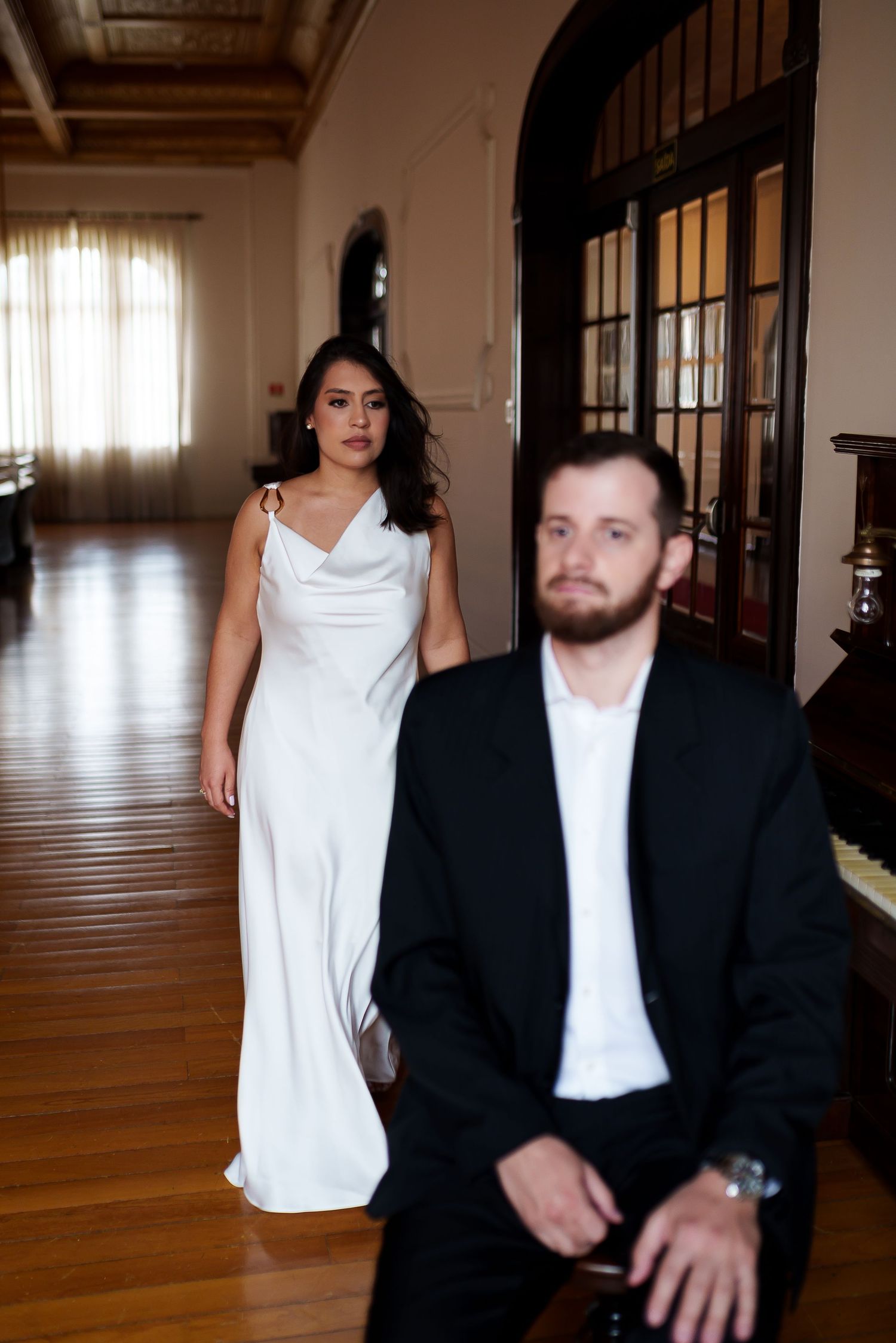 Bride in white satin gown walks toward groom in black suit inside elegant venue with hardwood floors.