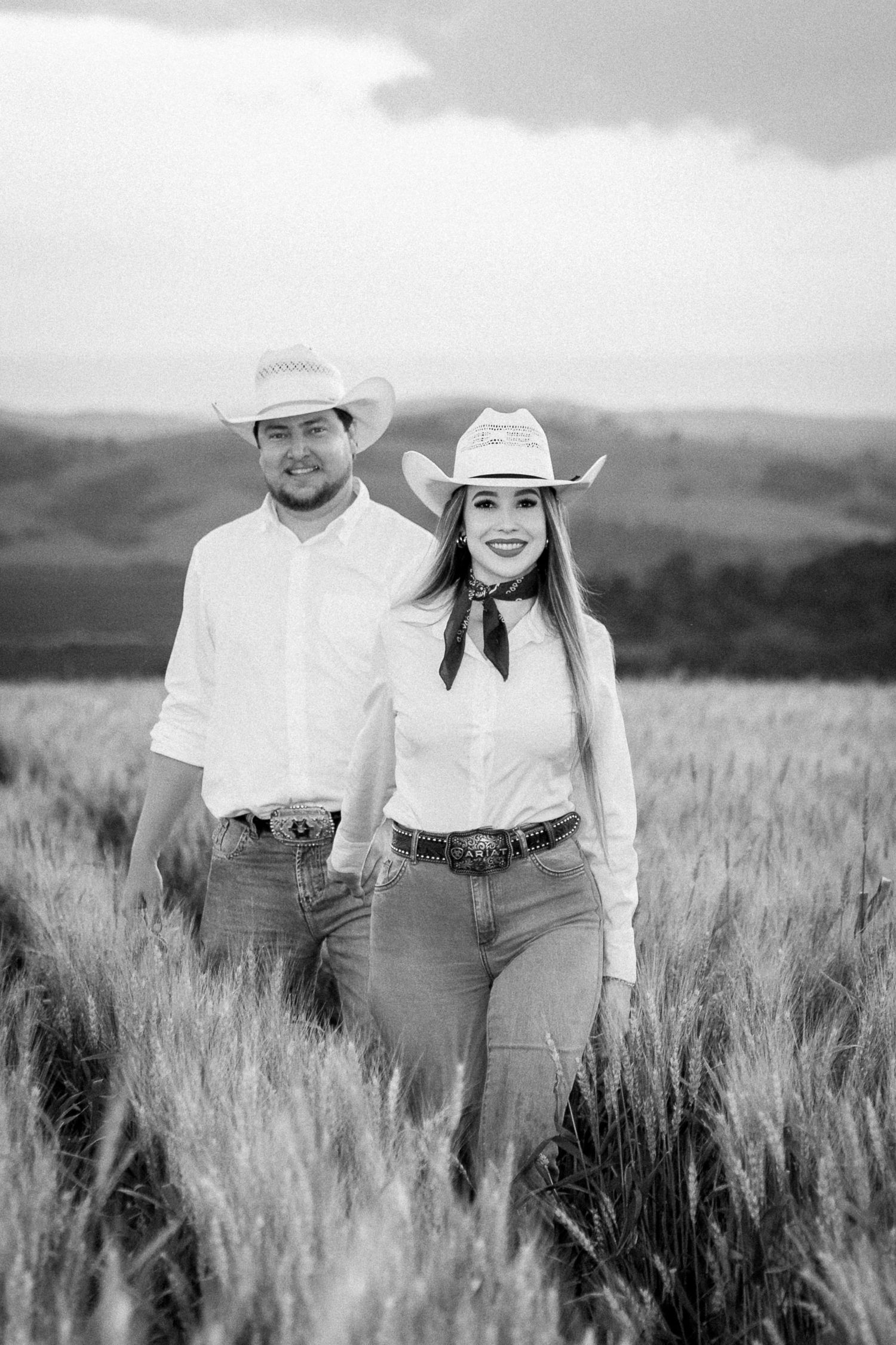 Black and white photo of a couple in cowboy hats and jeans posing in a wheat field with rolling hills behind them.