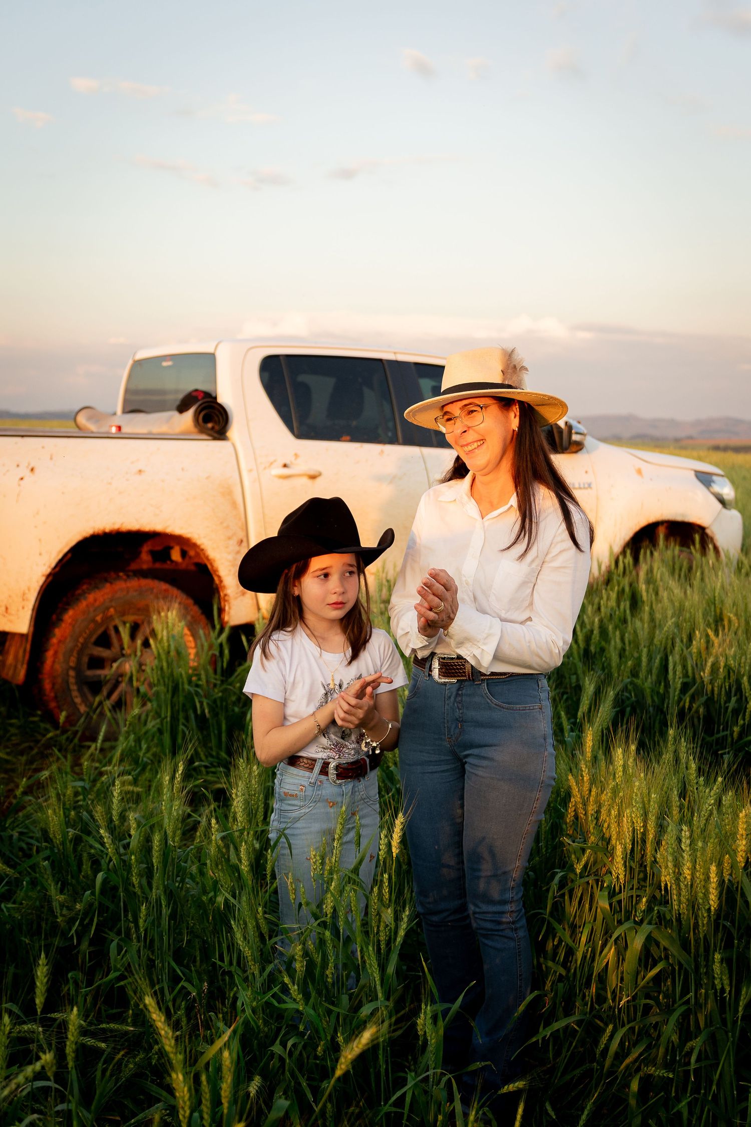 A woman and girl in cowboy hats stand in a green wheat field beside a muddy white pickup truck at golden hour.