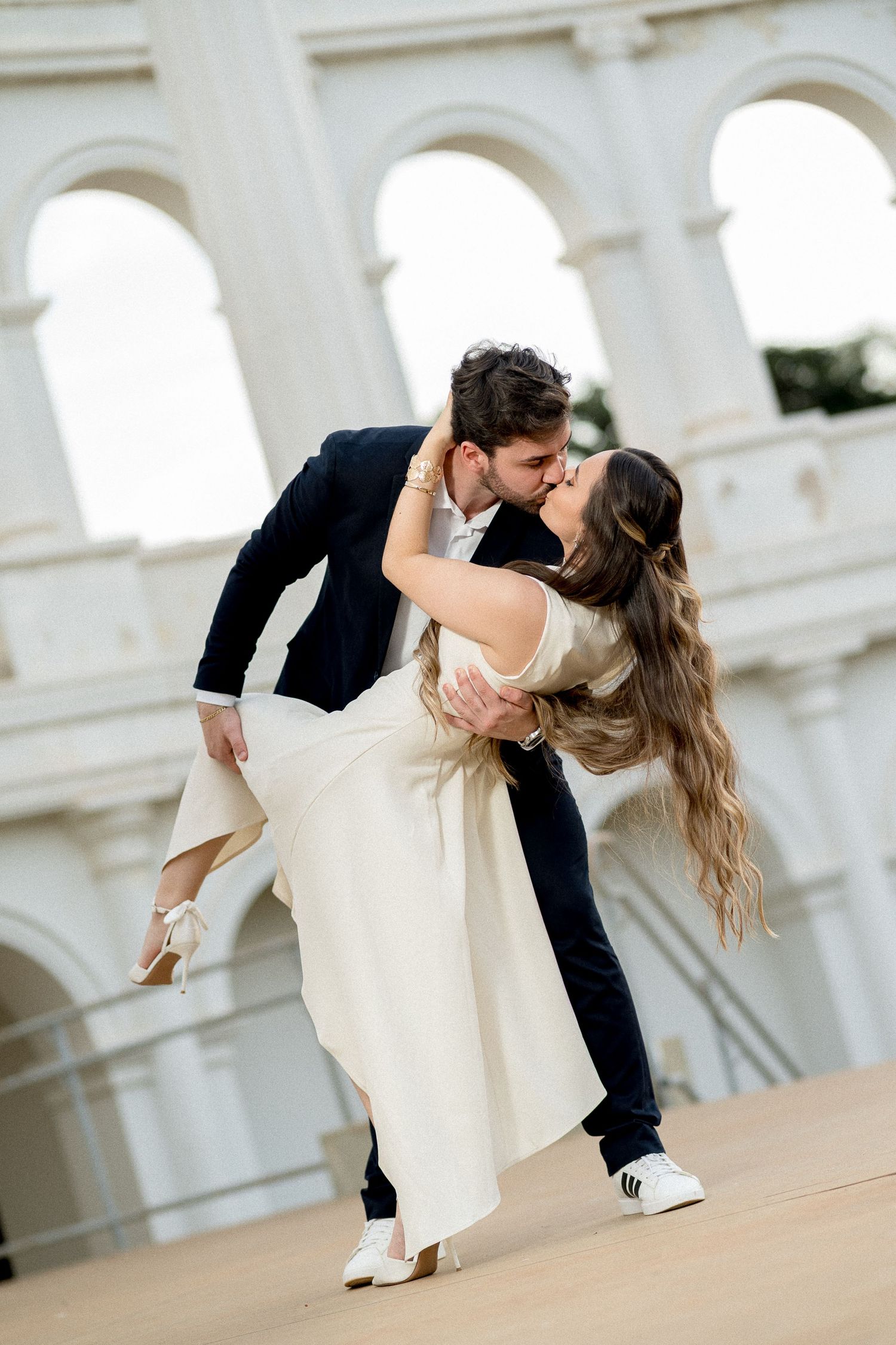 Couple sharing a romantic dip kiss in front of white arched building, woman in cream dress, man in dark suit.