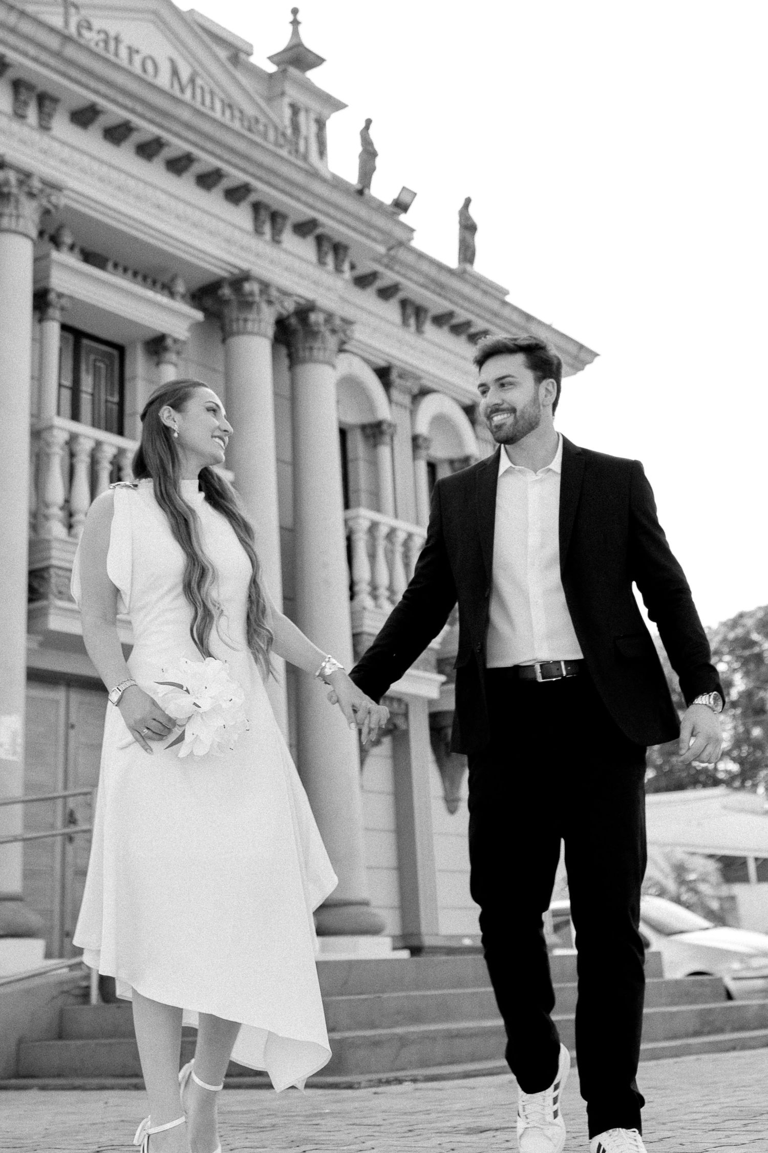 Black and white photo of a smiling couple in wedding attire holding hands in front of a grand classical building.