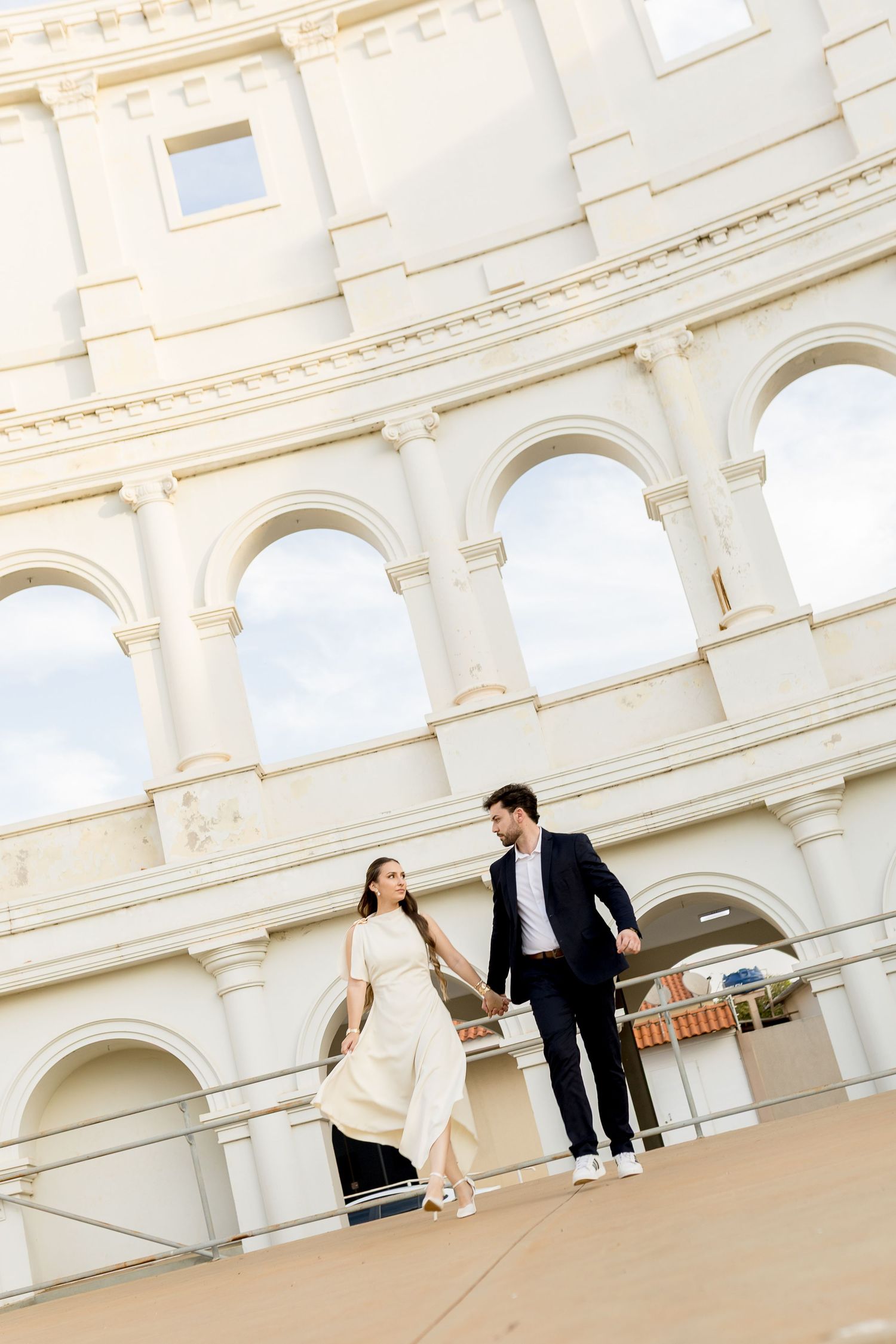 Couple holding hands walking in front of the Colosseum in Rome, woman in white dress, man in dark suit.
