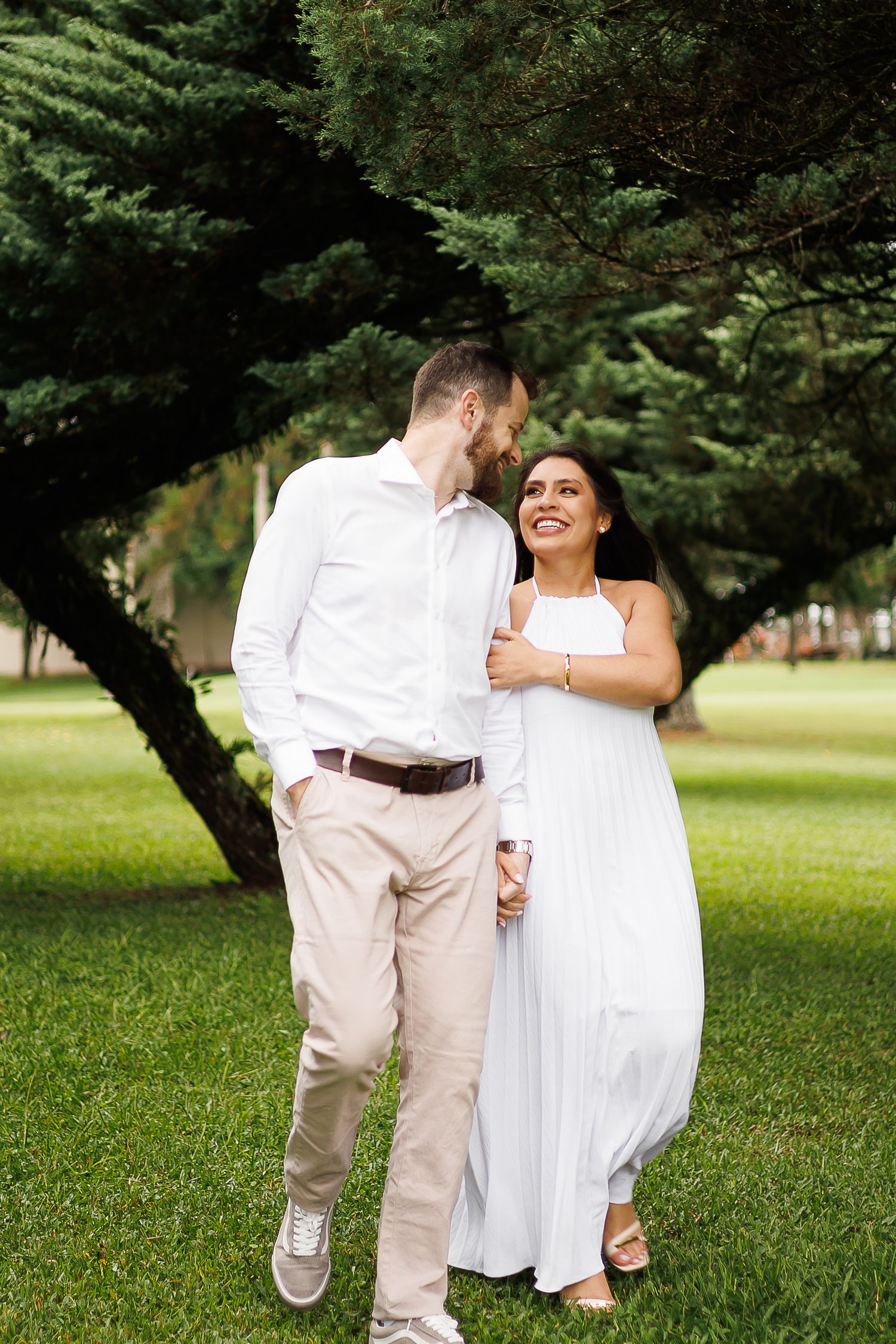 A couple in white attire share a romantic moment under a lush green tree in a park.